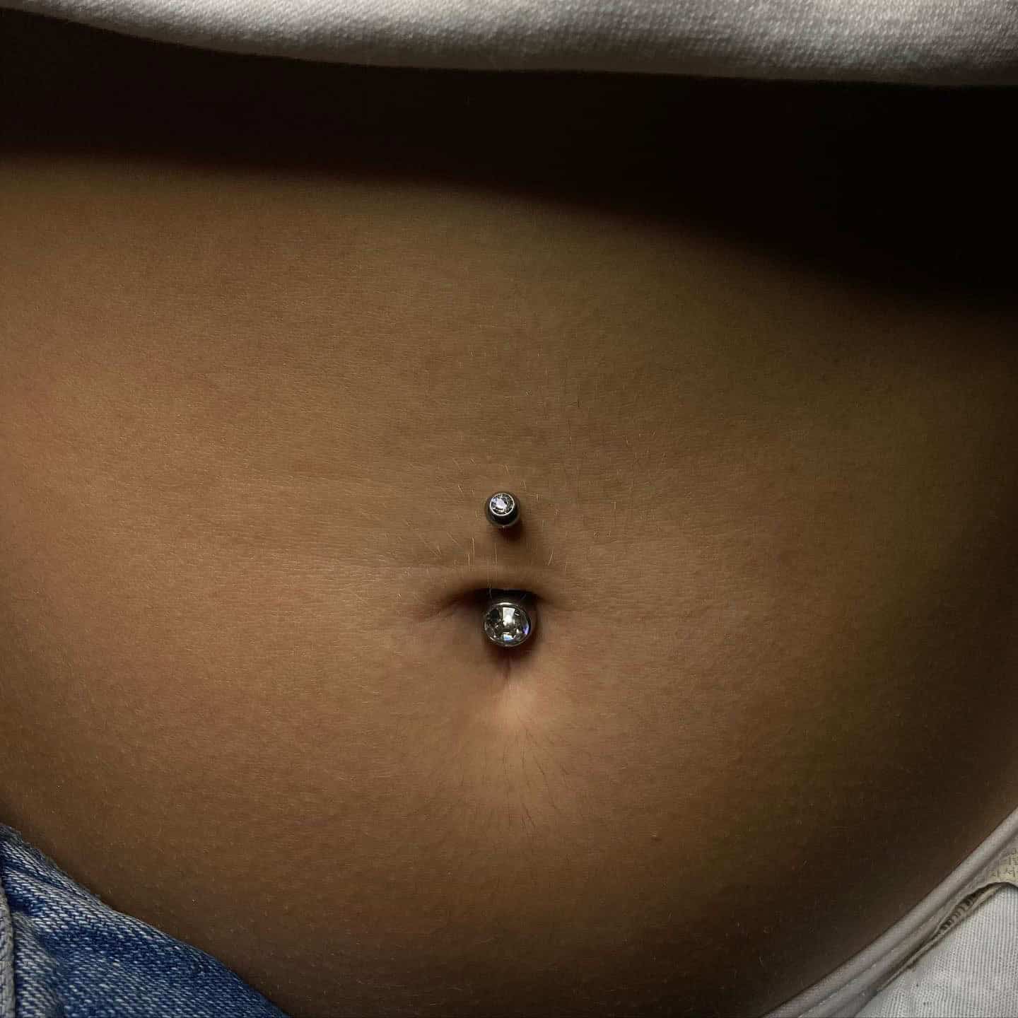 7sinstattoo.pl Zbliżenie pępka osoby ozdobionego piercingiem 01E732A3-9C82-4252-8736-0EEBB9E24FD4. Piercing ma srebrny ćwiek z przezroczystym kamieniem szlachetnym w górnej części i większą srebrną kulkę z przezroczystym kamieniem szlachetnym w dolnej części. Osoba ma na sobie jasny top i niebieskie dżinsy, idealne na wizytę w studio tatuażu Poznań. Najlepsze studio tatuażu Seven Sins Tattoo