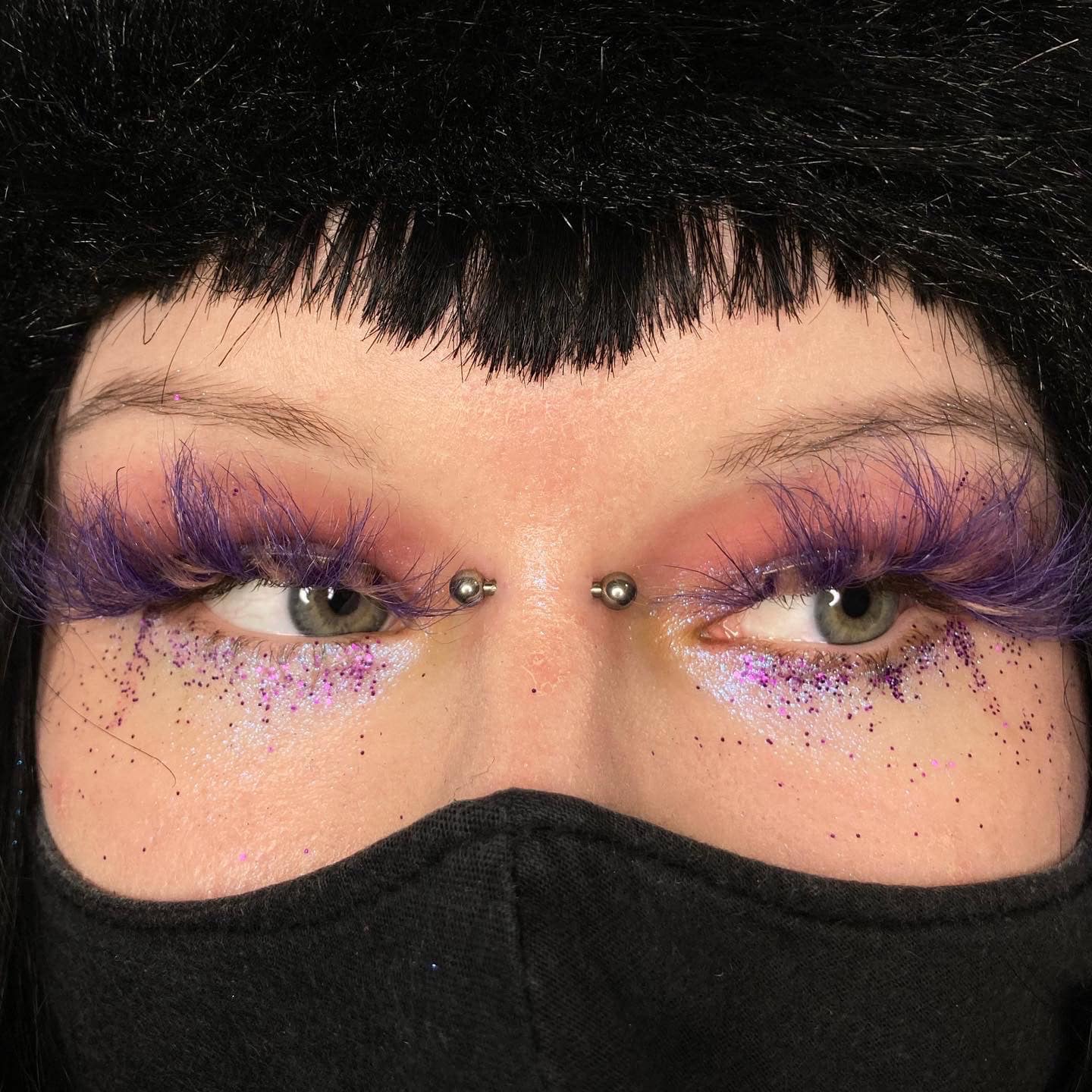 7sinstattoo.pl Zbliżenie ukazuje oczy osoby ozdobione dramatycznymi, puszystymi fioletowymi rzęsami i błyszczącym fioletowym brokatem. Ich metalowe kolczyki mostka dodają odważnego wyglądu, podkreślonego czarnym kapeluszem z grzywką i czarnym materiałem zakrywającym dolną część twarzy. Ta estetyka doskonale uosabia odważny styl często celebrowany w studio tatuażu Poznań. Cały wygląd zawiera elementy z produktu 058FBC45-9358-4B63-9A3E-73BD39EB5230. Najlepsze studio tatuażu Seven Sins Tattoo