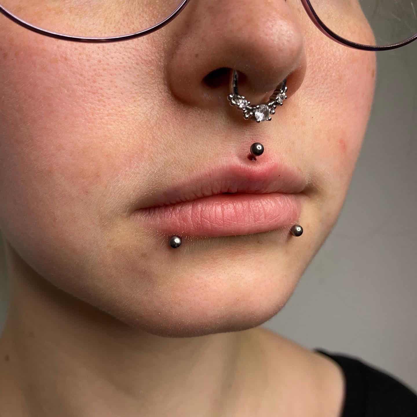 7sinstattoo.pl Zbliżenie dolnej części twarzy osoby, prezentujące jej misterne piercingi twarzy, zostało pięknie uchwycone w studio tatuażu Poznań. Osoba jest ozdobiona piercingiem przegrody nosowej o misternym wzorze i trzema piercingami warg: jednym umieszczonym centralnie pod dolną wargą i dwoma po obu stronach nad brodą. Nosi również okulary. Ten obraz jest zidentyfikowany jako Nazwa produktu 06219259-71A6-4A73-B889-7A84A896FAC3. Najlepsze studio tatuażu Seven Sins Tattoo