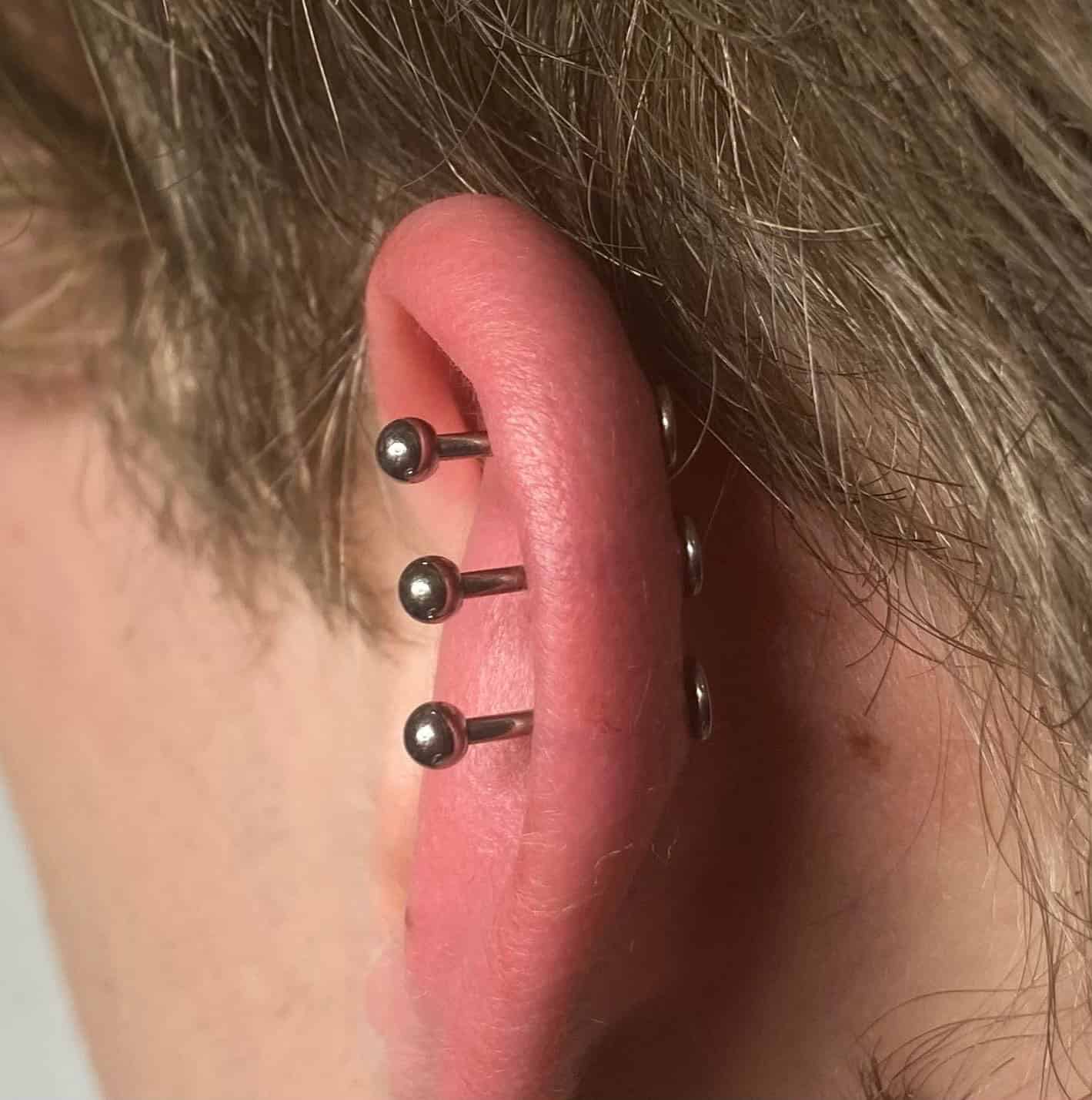 7sinstattoo.pl Zbliżenie ucha przekłutego trzema przemysłowymi sztangami 0B2BB22E-0D99-4030-A1D5-28F5857EDCF6, z których każda ma małe srebrne kulki na obu końcach. Ucho ma czerwonawe odcienie wokół przekłuć, na tle jasnobrązowych włosów. Zrobione w Studio Tatuaże Poznań, znanym z wyjątkowej dbałości o szczegóły. Najlepsze studio tatuażu Seven Sins Tattoo