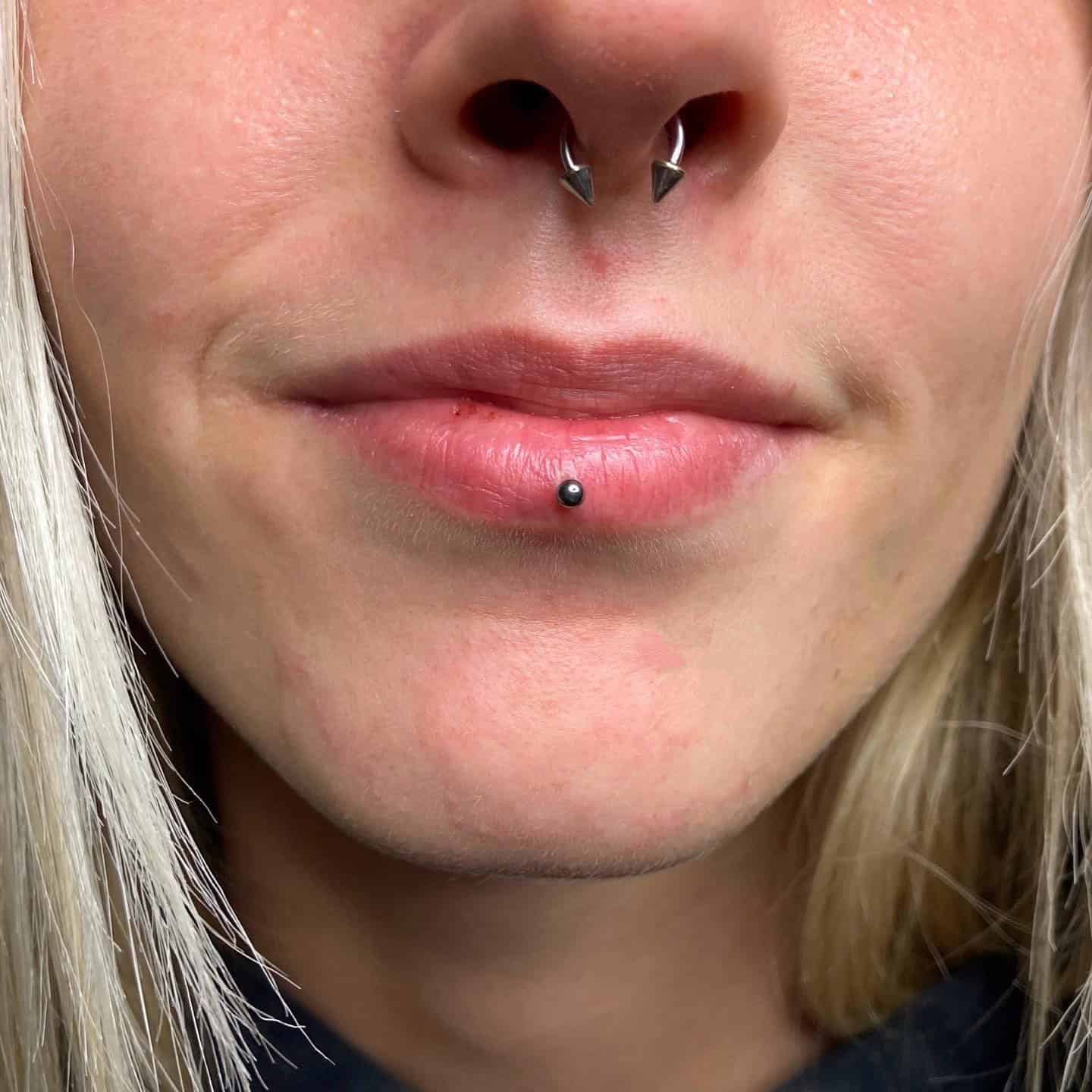 7sinstattoo.pl Zbliżenie ust i nosa osoby ukazuje piercing przegrody nosowej z podkową ozdobioną dwoma kolcami, obok piercingu wargi dolnej z małą kulką. Osoba ma jasną cerę i blond włosy, charakterystyczne dla klientów studia tatuażu Poznań, prezentującego produkt 16924757-9863-467A-A3B5-E3B45A85F898. Najlepsze studio tatuażu Seven Sins Tattoo