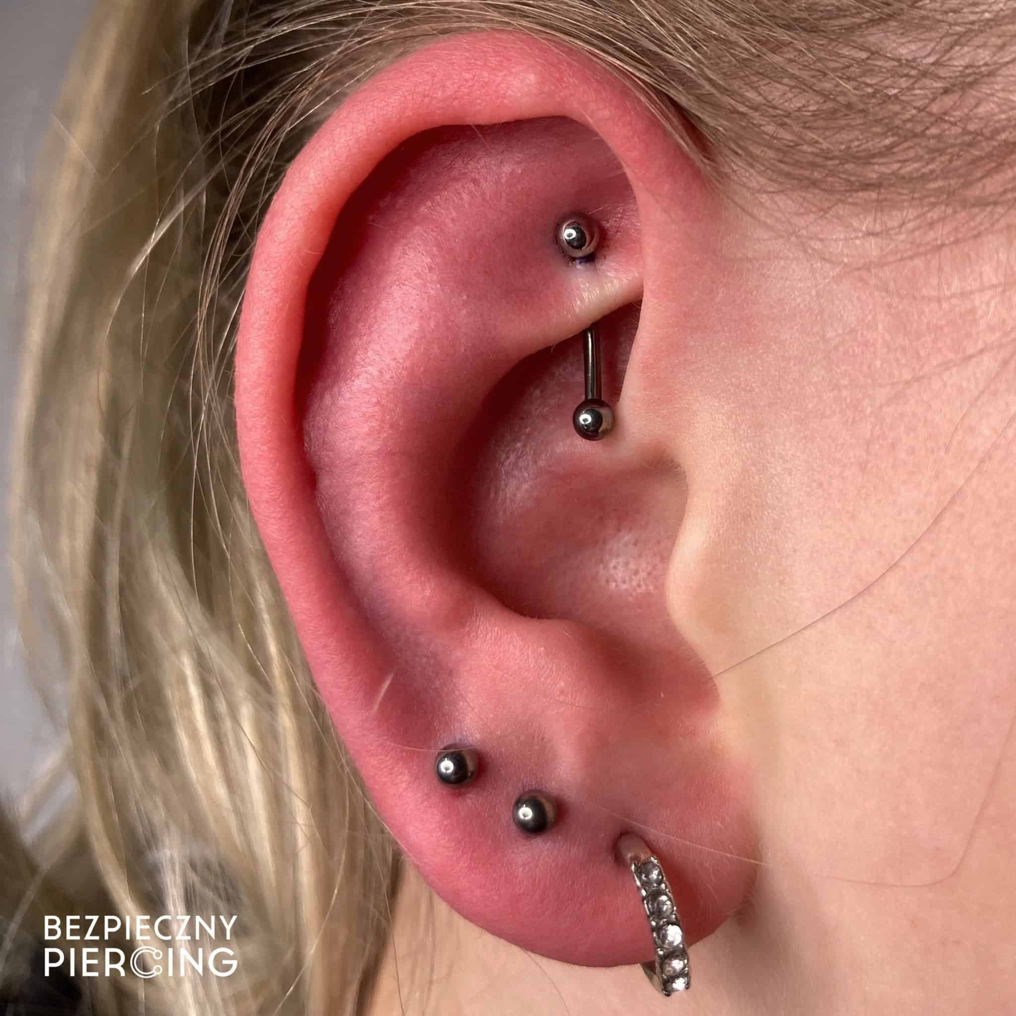 7sinstattoo.pl Zbliżenie ucha osoby ozdobionego trzema różnymi piercingami: sztangą w chrząstce, dwoma kolczykami w płatku i kółkiem wysadzanym kamieniami szlachetnymi w dolnym płatku. Obraz zawiera tekst w lewym dolnym rogu, który brzmi „BEZPIECZNY PIERCING” ze studia tatuaży Poznań. Polecany produkt: 17A15EDF-8872-4FA1-9B55-F0A3AE14C62B. Najlepsze studio tatuażu Seven Sins Tattoo