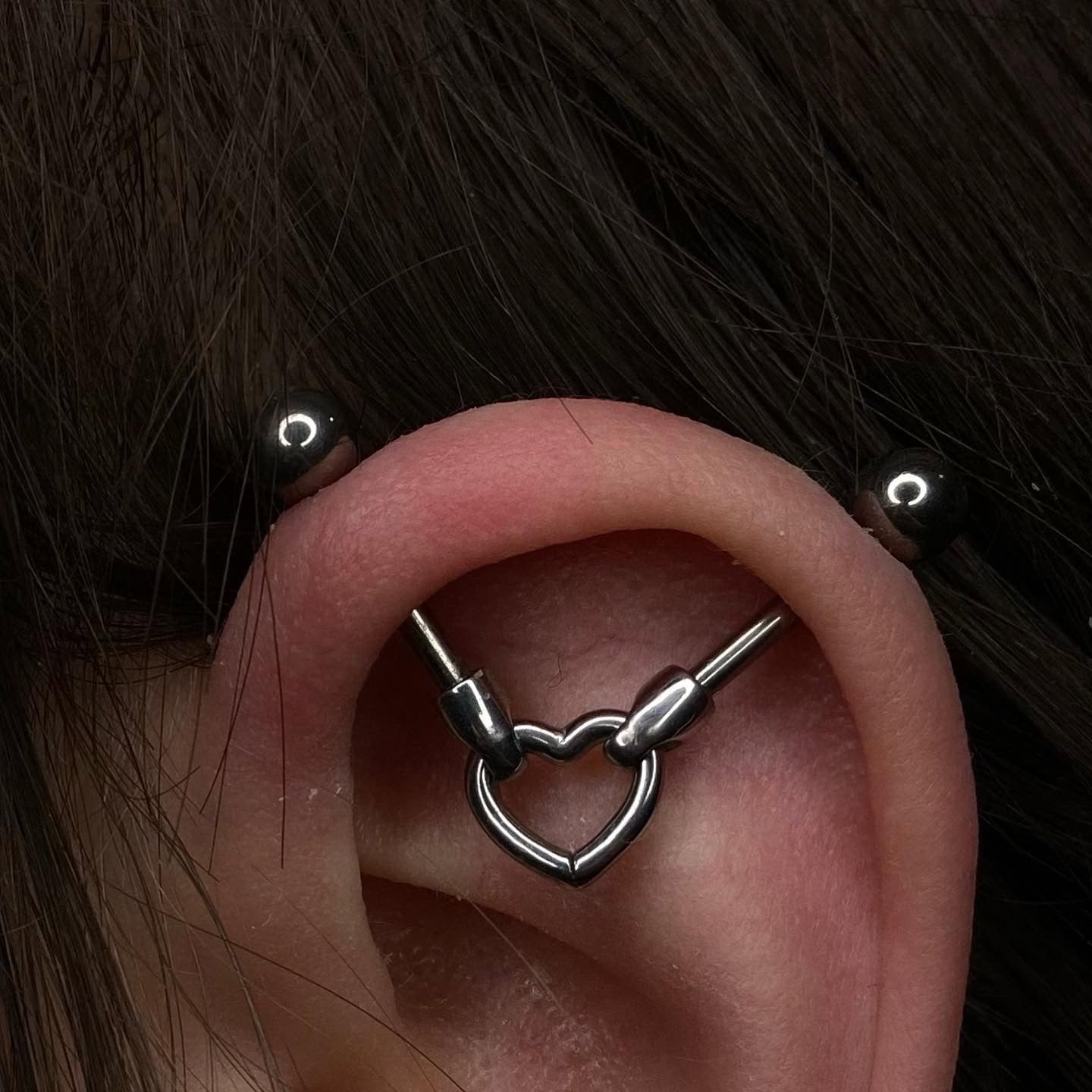 7sinstattoo.pl Zbliżenie ucha z industrialnym piercingiem. Kolczyk ma dwa końce, które łączą się z metalowym środkiem w kształcie serca na środku ucha. Biżuteria z produktu 1DCFB785-E9DE-49EC-84CC-3B1133C71388 jest osadzona na tle ciemnych włosów, eksponując misterne dzieło, które można znaleźć w studio tatuażu Poznań. Najlepsze studio tatuażu Seven Sins Tattoo