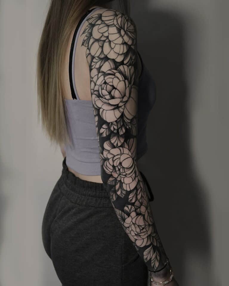 Kobieta z długimi włosami, ubrana w krótki szary top i ciemne spodnie dresowe, jest pokazana z tyłu i z boku. Na jej prawym ramieniu znajduje się szczegółowy czarno-szary rękaw z tatuażem kwiatowym. Neutralne tło podkreśla projekt, prezentując kunszt Seven Sins Tattoo Studio w Poznaniu.