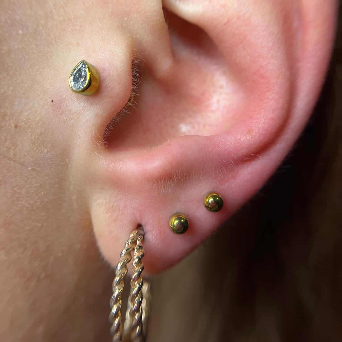 7sinstattoo.pl Zbliżenie ucha z trzema piercingami, zrobione w studio tatuażu Poznań. Na górnej części ucha znajduje się mały kolczyk z kamieniem szlachetnym, na dolnej dwa małe złote kolczyki w kształcie kulek, a na płatku ucha gruby, skręcony złoty kolczyk-kółko. Wszystkie prezentowane kolczyki pochodzą z produktu 2057CC28-B589-4B9C-9277-4E72C18439B6. Najlepsze studio tatuażu Seven Sins Tattoo