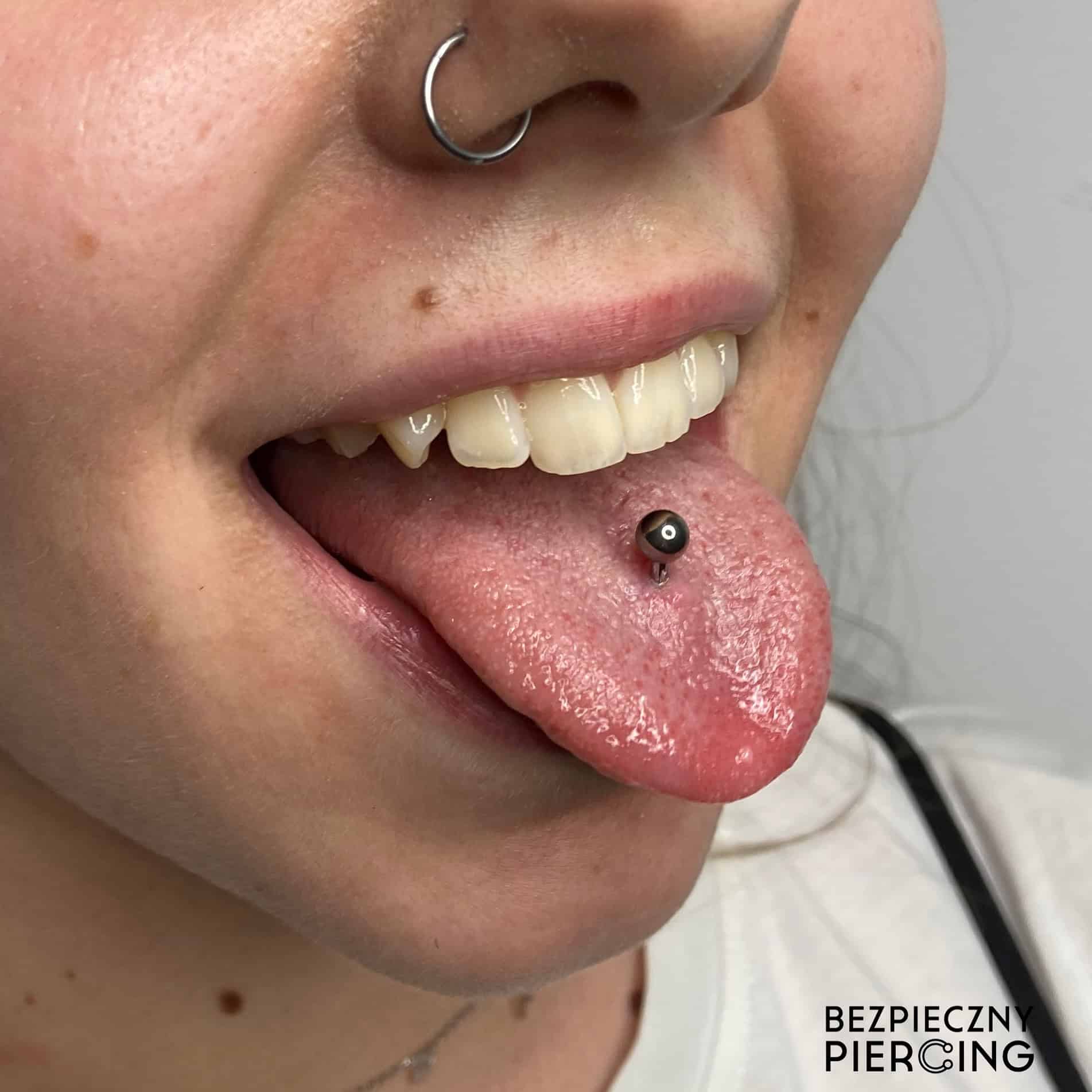 7sinstattoo.pl Zbliżenie twarzy osoby ukazuje kolczyk w lewym nozdrzu i piercing języka ozdobiony metalowym sztangą. Osoba uśmiecha się z otwartymi ustami, eksponując białe zęby. W prawym dolnym rogu tekst „BEZPIECZNY PIERCING” podkreśla kompetencje naszego poznańskiego studia tatuażu, prezentując produkt 23B5387E-E272-4E64-AB43-AE5070EC0043. Najlepsze studio tatuażu Seven Sins Tattoo