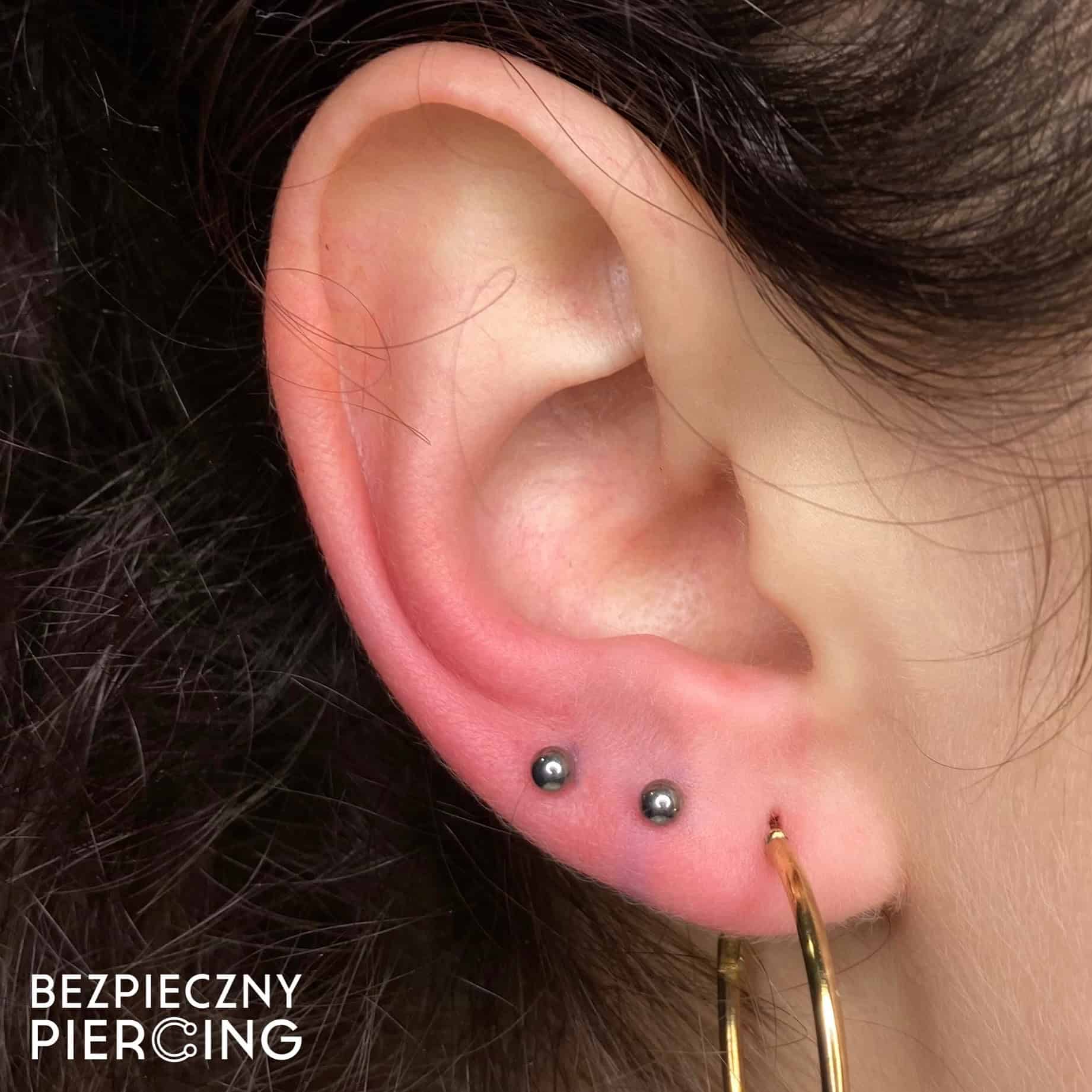 7sinstattoo.pl Zbliżenie na ucho osoby z dwoma małymi czarnymi kolczykami na płatku i dużym złotym kolczykiem w kształcie koła w dolnym płatku. W tle widać ciemne włosy, a w lewym dolnym rogu wyświetlany jest tekst „BEZPIECZNY PIERCING”, podświetlający produkt 27664927-1D55-4BE8-BE0E-260FE4AFE6BE z naszego studia tatuaży Poznań. Najlepsze studio tatuażu Seven Sins Tattoo