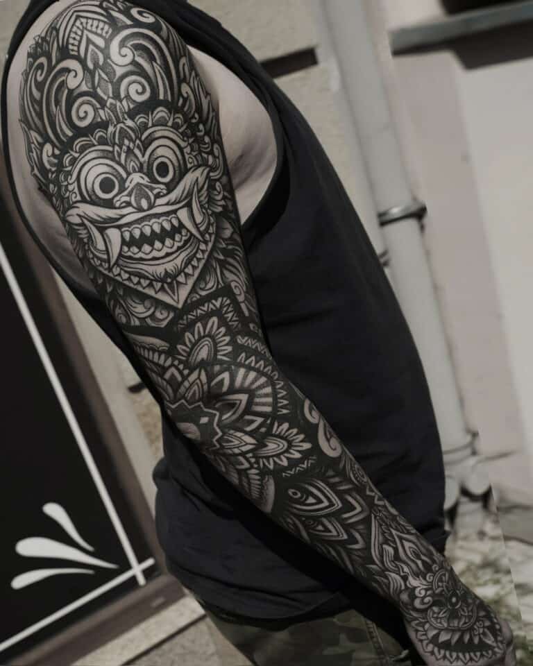 Osoba z pełnym tatuażem na prawym ramieniu, prezentująca misterne czarno-szare wzory przedstawiające stylizowaną twarz z dużymi oczami i otwartymi ustami, otoczoną geometrycznymi i kwiatowymi wzorami. Ma na sobie czarny top bez rękawów i spodnie moro, stoi na zewnątrz przy ścianie z widocznymi rurami. To oszałamiające dzieło sztuki powstało w studiu Seven Sins Tattoo w Poznaniu.