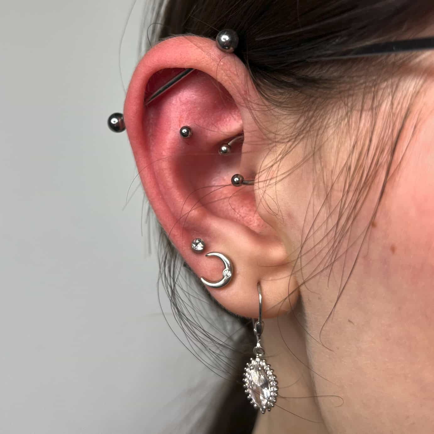 7sinstattoo.pl Zbliżenie ucha osoby ozdobionego wieloma piercingami w najlepszym studiu tatuażu w Poznaniu. Ucho ma mieszankę biżuterii, w tym kolczyki, małe kółko z zawieszką w kształcie półksiężyca i zwisający kolczyk z ozdobnym wzorem i dużym, przezroczystym kamieniem szlachetnym z kolekcji **32729F66-76D4-4F28-8B98-382AE417F714**. Włosy osoby są schowane za uchem. Najlepsze studio tatuażu Seven Sins Tattoo