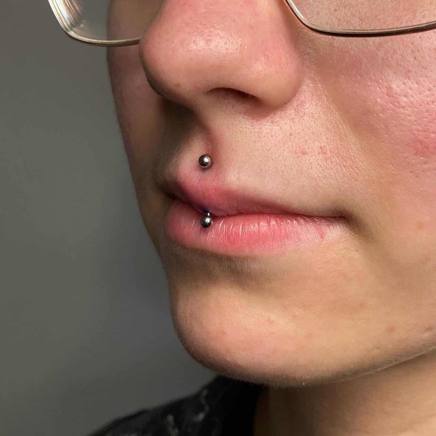 7sinstattoo.pl Zbliżenie twarzy osoby noszącej okulary, z pionowym piercingiem wargi górnej ozdobionym jednym koralikiem nad górną wargą i drugim pod dolną wargą ze studia tatuażu Poznań. Tło jest gładkie i nieostre. Produkt użyty do piercingu jest zidentyfikowany jako 3CCABBAA-8141-4D1B-947C-B3DD1DF3D99F. Najlepsze studio tatuażu Seven Sins Tattoo