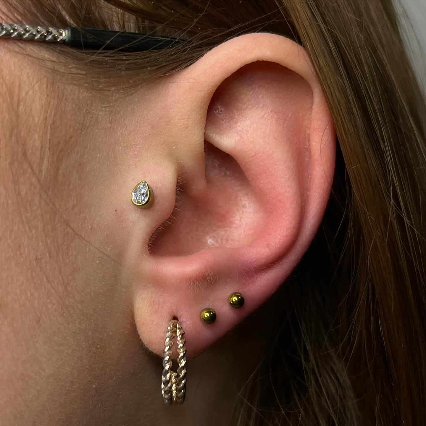 7sinstattoo.pl Zbliżenie ucha osoby pokazujące różne kolczyki, w tym produkt 409414AB-0BA3-4CBA-9D6A-3D1B33ECAE16. W górnej chrząstce znajduje się gruszkowaty diamentowy kolczyk, dwa małe złote kolczyki wzdłuż zewnętrznej części ucha i teksturowana złota obręcz w płatku ucha. Osoba nosi również okulary z czarno-złotymi oprawkami, stylowo uchwycone w study tatuażu Poznań. Najlepsze studio tatuażu Seven Sins Tattoo