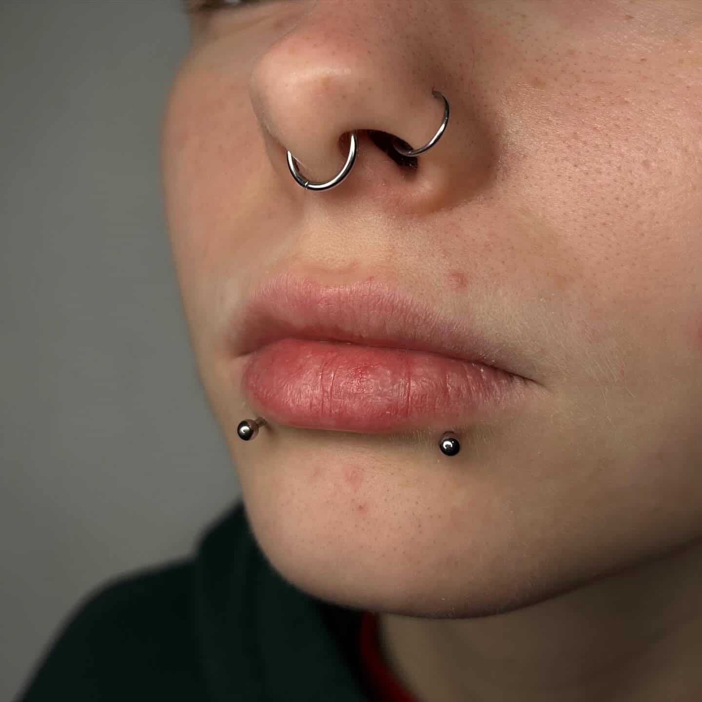 7sinstattoo.pl Zbliżenie dolnej części twarzy osoby pokazujące dwa piercingi wargi z małymi, czarnymi koralikami po obu stronach dolnej wargi i piercing przegrody nosowej z kółkiem w kształcie podkowy. Osoba ma neutralny wyraz twarzy, a część ciemnej koszuli jest widoczna. Zrobione w jednym z najlepszych studiów tatuaży w Poznaniu. Prezentuje produkt 410E2558-F30F-417D-B386-375B79E61F1C. Najlepsze studio tatuażu Seven Sins Tattoo