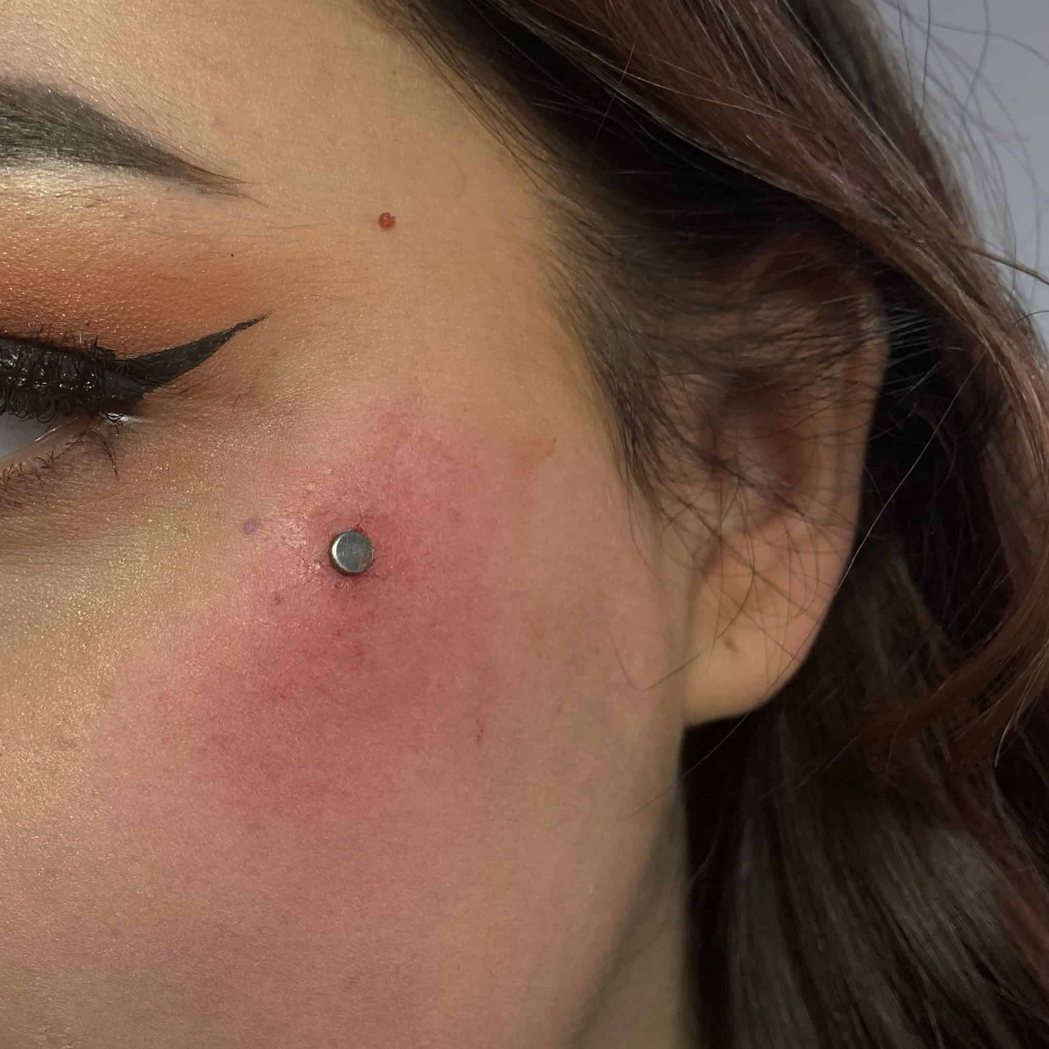 7sinstattoo.pl Zbliżenie lewej strony twarzy kobiety, prezentujące świeży piercing policzka z okrągłym, srebrnym ćwiekiem ozdobionym produktem 4468FFAC-CA96-4AEA-966D-A3FCD9115568. Przekłuty obszar wydaje się lekko zaczerwieniony i opuchnięty. Ma skrzydlatą kreskę na powiece i lekko nałożony makijaż, a jej ciemnobrązowe włosy są częściowo widoczne — doskonały przykład z najlepszego studia tatuażu Poznań. Najlepsze studio tatuażu Seven Sins Tattoo