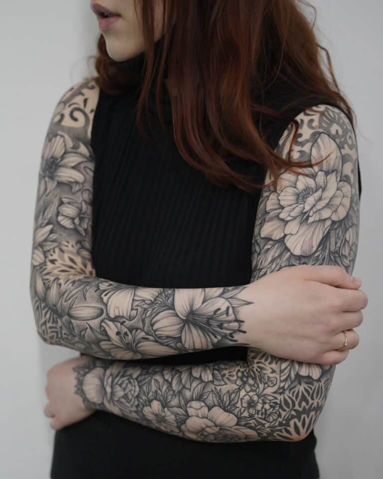Kobieta z długimi, rudymi włosami od ramion w dół, ubrana w czarny top bez rękawów. Obie jej ręce są skrzyżowane i całkowicie pokryte szczegółowymi kwiatowymi tatuażami ze studia Seven Sins Tattoo w Poznaniu. Skomplikowane czarno-białe wzory przedstawiają różne kwiaty i liście rozciągające się od jej ramion do nadgarstków. Nosi również delikatny pierścionek na lewej ręce.