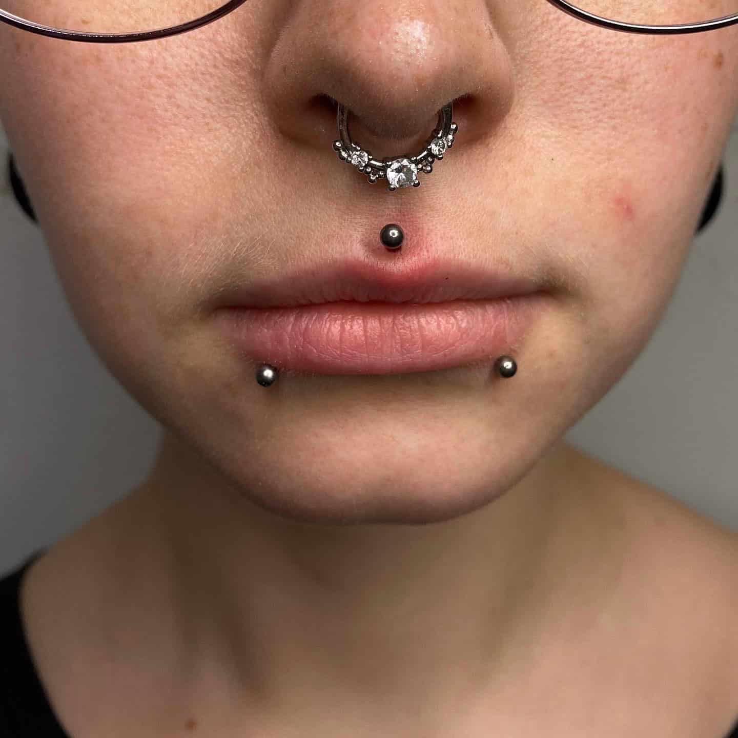 7sinstattoo.pl Zbliżenie twarzy osoby z wieloma piercingami w studio tatuażu Poznań. Są to piercing przegrody nosowej z biżuterią, pionowy piercing wargi z kulką na końcu oraz dwa piercingi po każdej stronie dolnej wargi. Osoba ma na sobie okulary, a część jej szyi i koszuli jest widoczna. Nazwa produktu: 6107C23D-E30B-4A0D-80A2-0091C4B26C7C. Najlepsze studio tatuażu Seven Sins Tattoo