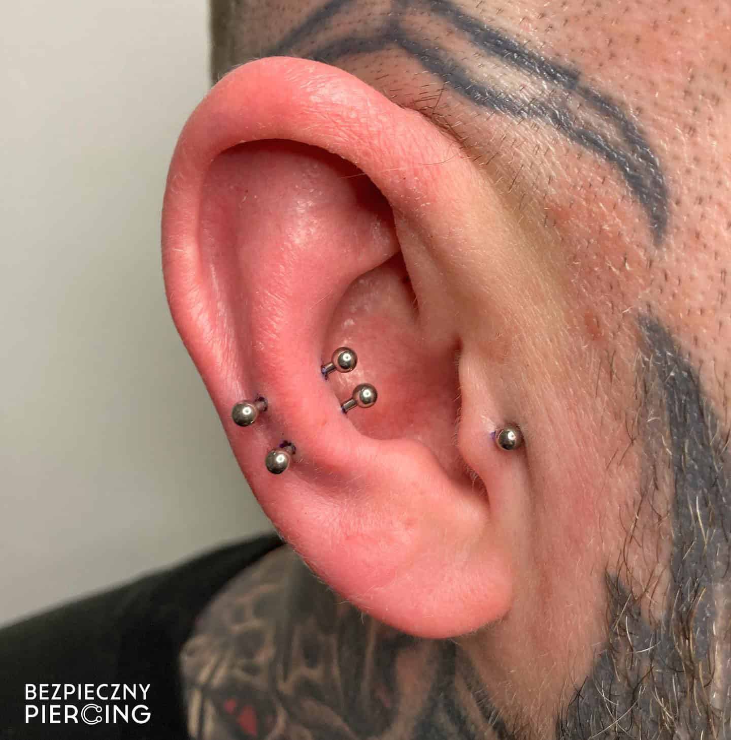 7sinstattoo.pl Zbliżenie ucha osoby ozdobionego wieloma piercingami, z dwoma srebrnymi sztangami w wewnętrznej chrząstce i dwoma w zewnętrznej chrząstce. Tatuaże obejmujące ucho podkreślają kompetencje studia tatuażu Poznań. Tekst „BEZPIECZNY PIERCING” jest wyświetlany w lewym dolnym rogu. Nazwa produktu: 610D03E3-B222-4A7B-A344-19391237608F. Najlepsze studio tatuażu Seven Sins Tattoo