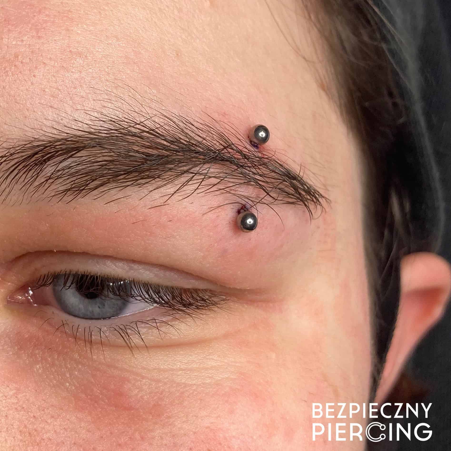 7sinstattoo.pl Zbliżenie brwi osoby ze świeżym poziomym piercingiem z dwoma czarnymi koralikami na każdym końcu. Skóra wokół piercingu jest lekko zaczerwieniona. Tekst „BEZPIECZNY PIERCING” jest widoczny w prawym dolnym rogu, podkreślając kompetencje najlepszego studia tatuażu Poznań i prezentując ich produkt, 624DB3A9-BF17-4CB5-8ADA-3FE1C3E632F1. Najlepsze studio tatuażu Seven Sins Tattoo