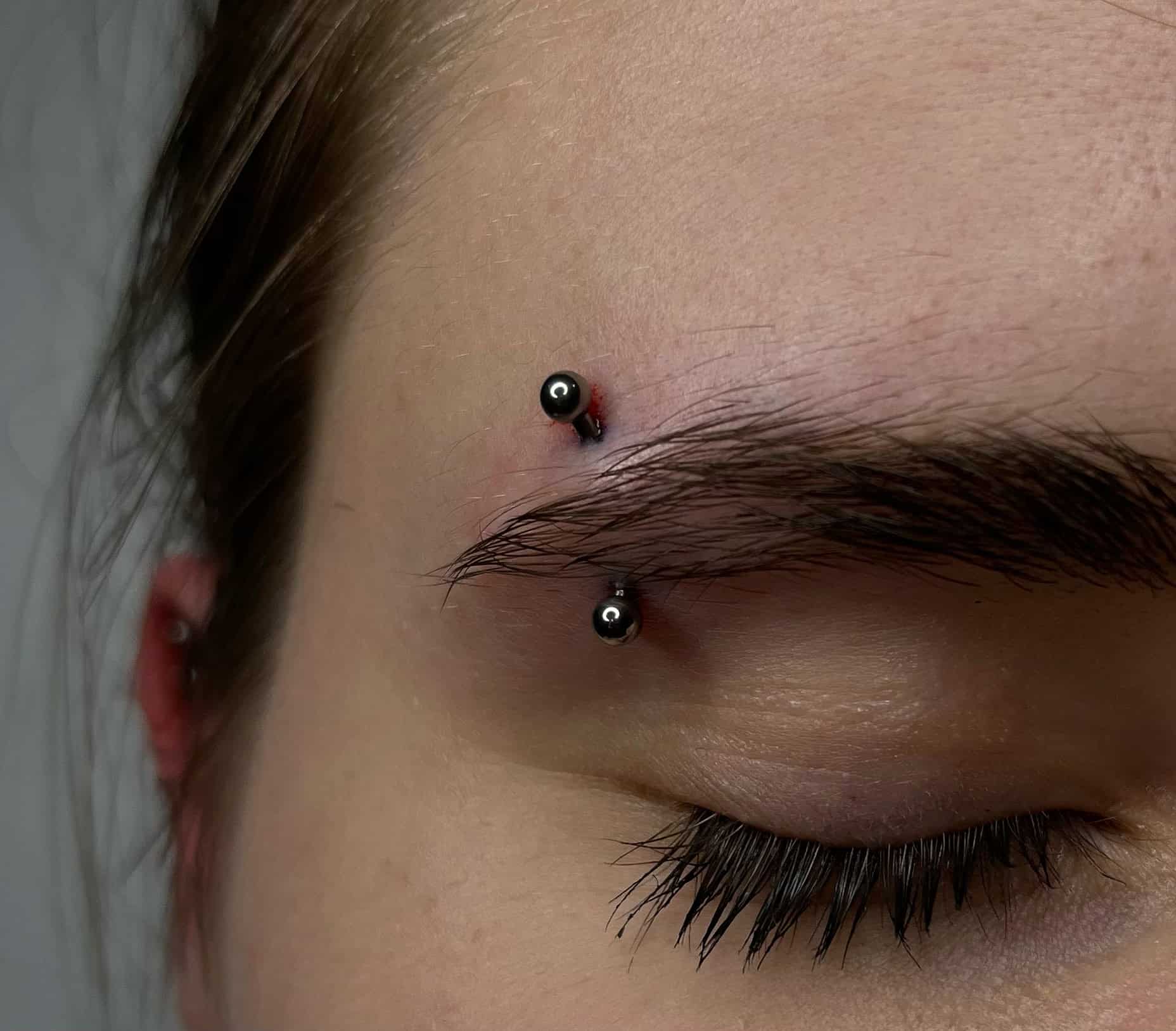 7sinstattoo.pl Zbliżenie na zamknięte oko osoby pokazujące piercing brwi. Piercing składa się z pręta z czarnymi kulkami na każdym końcu, przewleczonego przez skórę nad brwią, z odrobiną zaczerwienienia wokół punktów wejścia i wyjścia. Uchwycone w *studio tatuażu Poznań*, oko i rzęsy są widoczne. Polecany produkt: 62A1954A-9358-4D49-AA56-91AC6E07C17E. Najlepsze studio tatuażu Seven Sins Tattoo