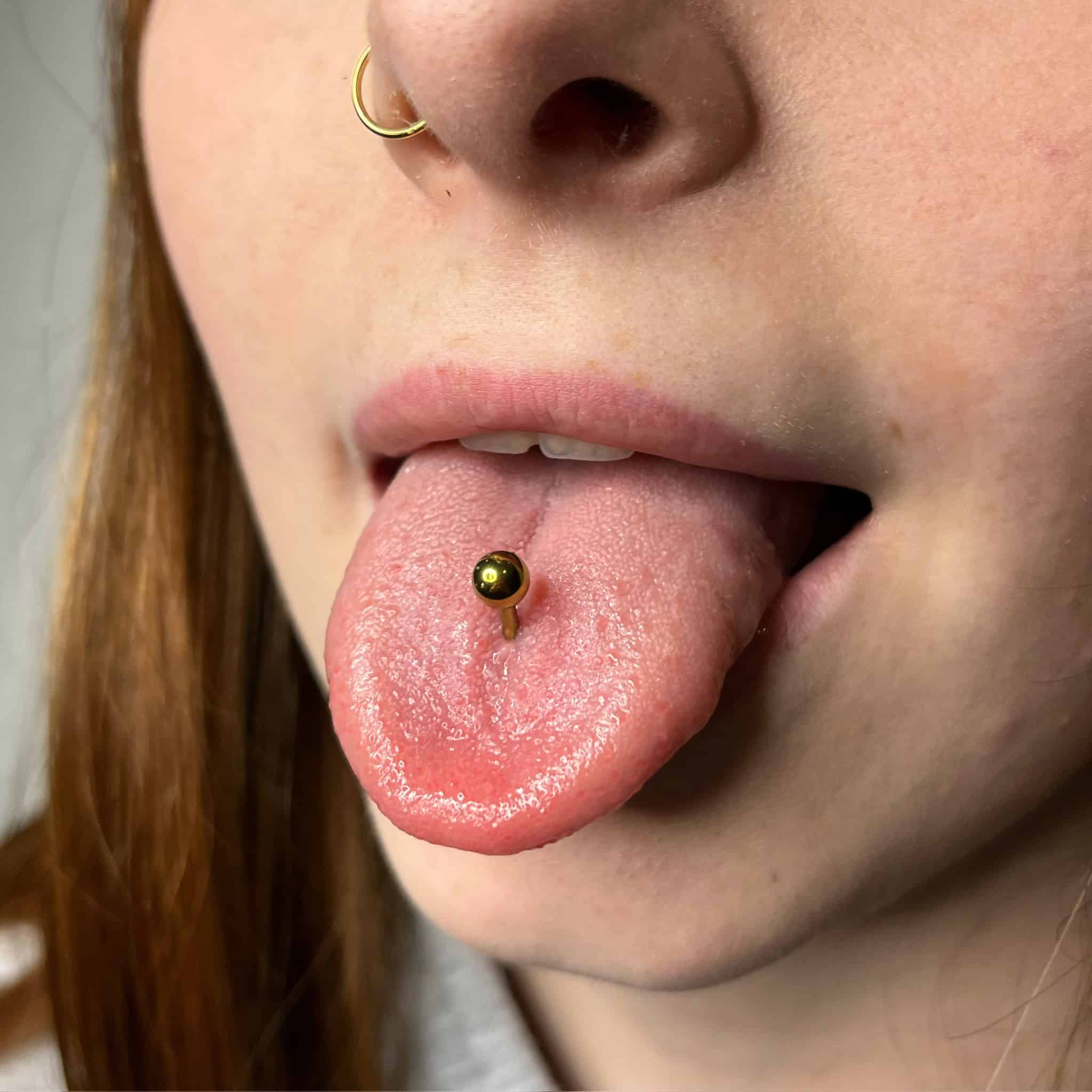 7sinstattoo.pl Zbliżenie przedstawia osobę o rudych włosach i jasnej karnacji, prezentującą złoty metalowy kolczyk do języka z produktu 698B21FB-BA40-494D-BB2C-A0EEFF19225A z najlepszego studia tatuażu Poznań. Dodatkowo ma pasujący złoty kolczyk do nosa i wystawia język, aby pokazać piercing. Najlepsze studio tatuażu Seven Sins Tattoo