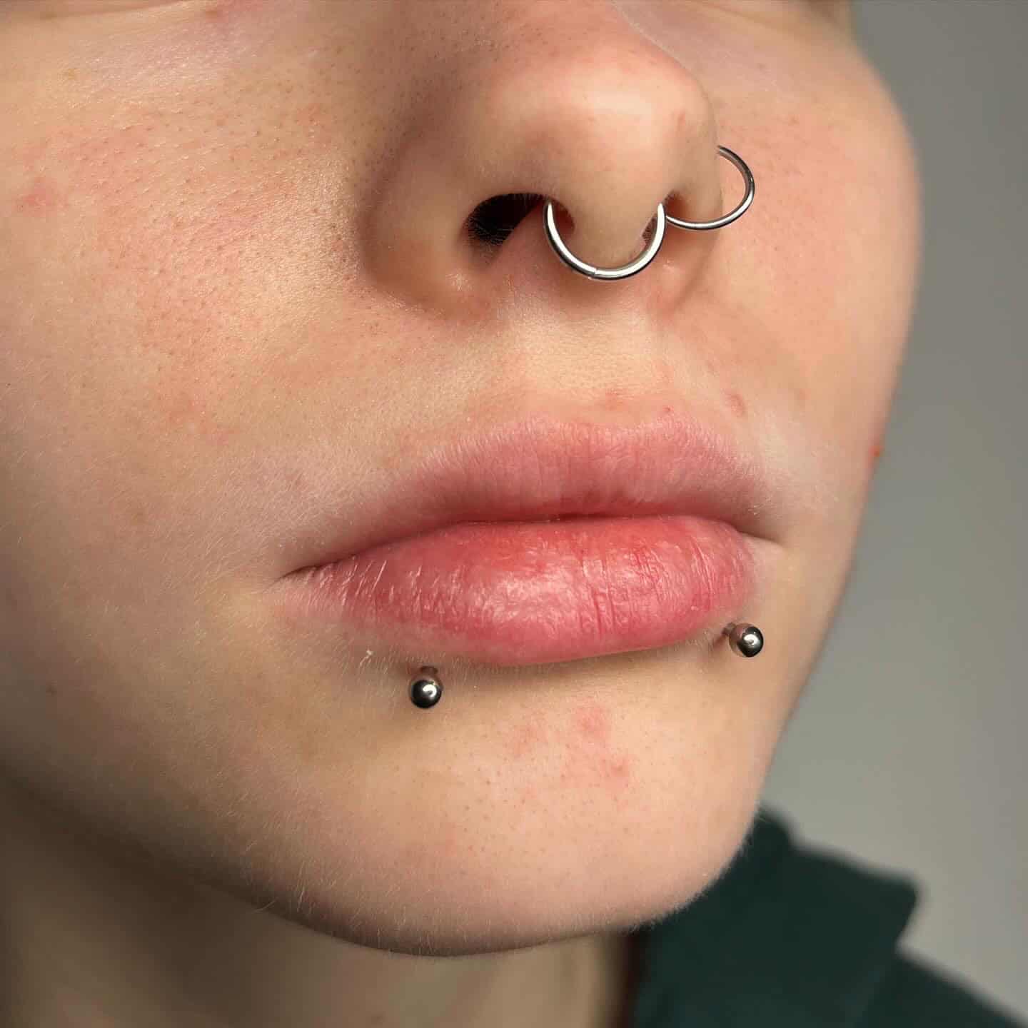 7sinstattoo.pl Zbliżenie nosa i ust osoby, z dwoma srebrnymi pierścieniami przebijającymi przegrodę nosową i dwoma kolczykami z kulkami zdobiącymi dolną wargę. Skóra wydaje się lekko zarumieniona na neutralnym tle, prezentując misterne wykonanie studia tatuażu Poznań dla produktu 6AED672E-4427-4355-9726-21B278396364. Najlepsze studio tatuażu Seven Sins Tattoo