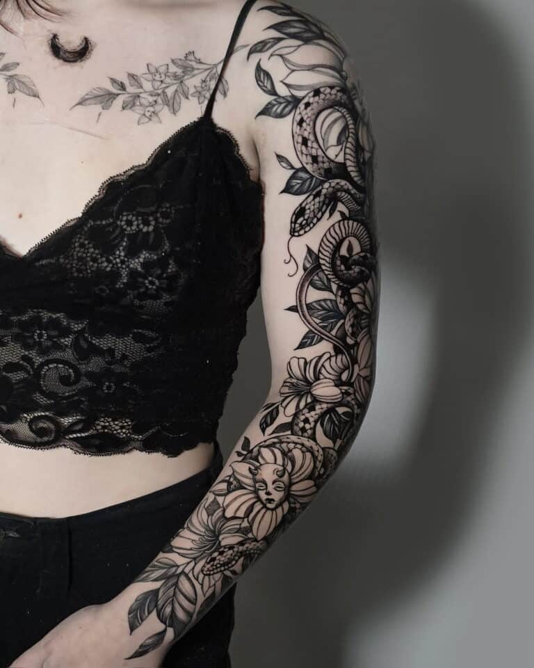 Osoba ubrana w misternie zaprojektowany czarny koronkowy top „7” i czarne spodnie z 7sinstattoo.pl prezentuje szczegółowy tatuaż na lewym ramieniu. Tatuaż, wykonany w Seven Sins Tattoo Studio w Poznaniu, zawiera różne motywy kwiatowe, płynne wzory, półksiężyc na ramieniu i twarz kobiety płynnie zintegrowaną z dziełem sztuki. Dodatkowe tatuaże są również widoczne na klatce piersiowej.