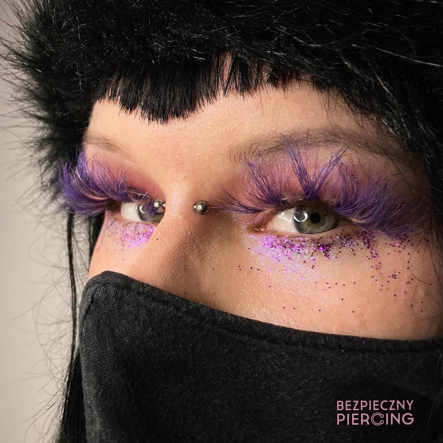7sinstattoo.pl Zbliżenie oczu osoby z misternymi fioletowymi, pierzastymi rzęsami i błyszczącym różowym cieniem do powiek, prezentujące mały metalowy piercing skóry nad grzbietem nosa. Nosi czarną maskę na twarzy i ma czarne włosy z grzywką. Tekst w prawym dolnym rogu mówi „BEZPIECZNY PIERCING - studio tatuaży Poznań”. Nazwa produktu: 79E5932F-E40F-48CF-A590-764DA0B0FDF1. Najlepsze studio tatuażu Seven Sins Tattoo