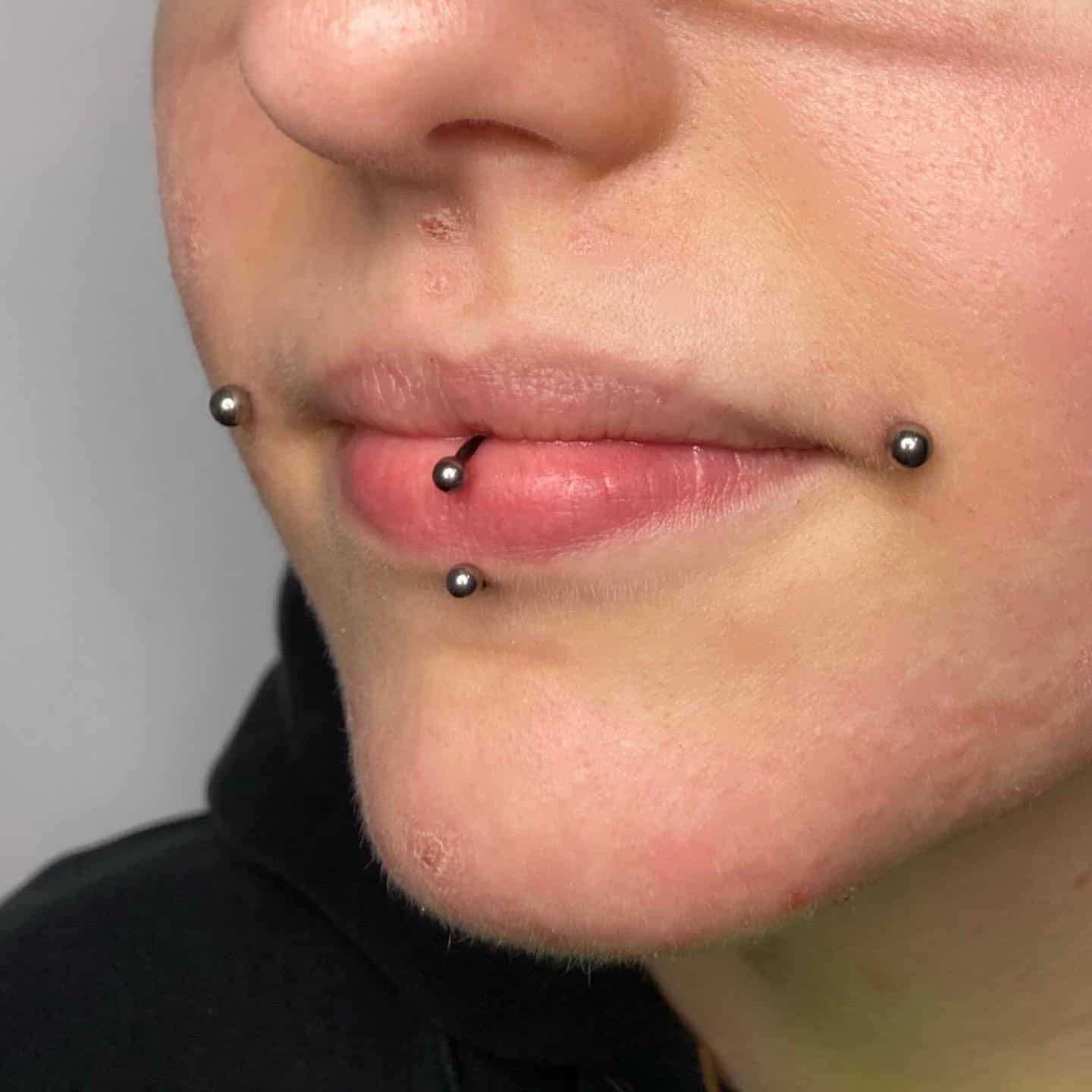 7sinstattoo.pl Zbliżenie dolnej części twarzy osoby z trzema piercingami w pobliżu ust: dwa po każdej stronie dolnej wargi (piercingi typu snake bite) i jeden piercing w środku. Metalowe ćwieki łapią światło, gdy noszą czarny top, prawdopodobnie marki 8A2DC004-DD18-4575-B6CF-AB860A969D5C. Najlepsze studio tatuażu Seven Sins Tattoo