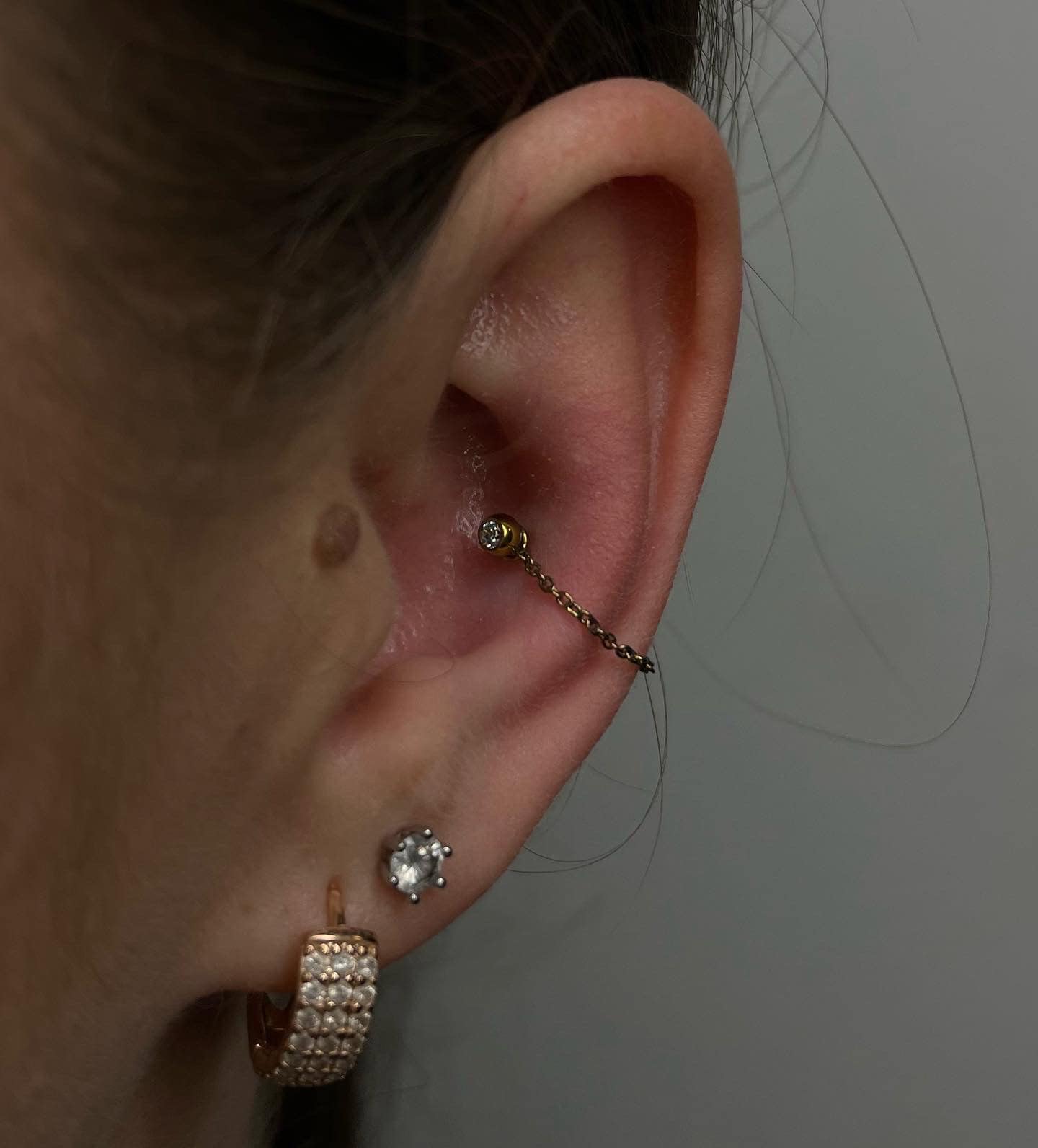 7sinstattoo.pl Zbliżenie na ucho z kilkoma piercingami wykonanymi w najlepszym studiu tatuażu w Poznaniu. W uchu znajduje się piercing daith ozdobiony produktem 923914D9-67C9-4504-BC07-900D423CC528, złotym kolczykiem w kształcie kwiatu połączonym cienkim łańcuszkiem z piercingiem helix. Dodatkowo są dwa piercingi płatka ucha: jeden z błyszczącym diamentowym kolczykiem, a drugi z kolczykiem w kształcie kółka. Najlepsze studio tatuażu Seven Sins Tattoo