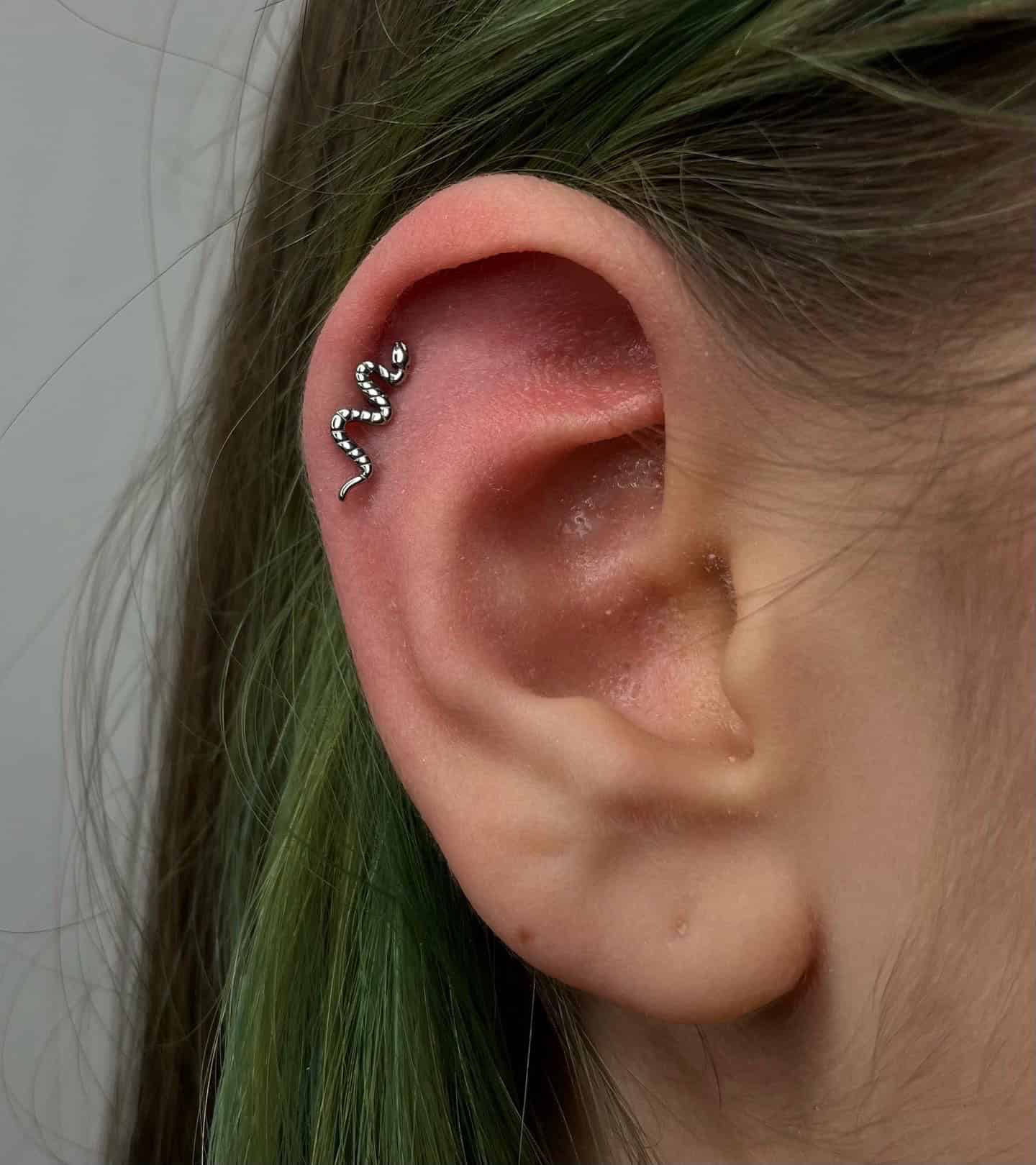 7sinstattoo.pl Zbliżenie ucha z włosami w odcieniu zieleni, z kolczykiem w kształcie węża w górnej chrząstce. Srebrny kolczyk o nazwie 9556375F-F7B9-4AF0-B339-33D35431B21B jest ozdobiony małymi kamieniami szlachetnymi, co nadaje mu lśniący wygląd. Miłośnicy sztuki na ciele powinni sprawdzić najlepsze studio tatuażu Poznań, aby znaleźć najwyższej klasy wzory. Najlepsze studio tatuażu Seven Sins Tattoo
