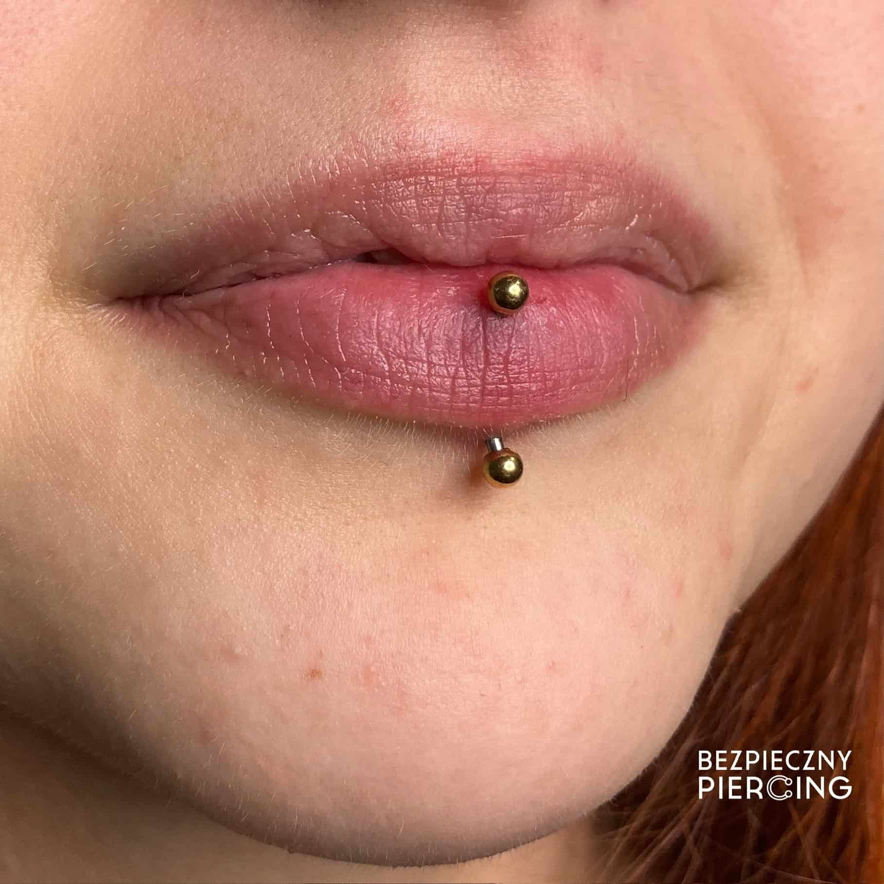 7sinstattoo.pl Zbliżenie ust osoby z pionowym piercingiem labret z tatuaż Poznań. Piercing przedstawia "A78660F6-9590-489D-8431-C9EE2E017FA6" z dwoma złotymi kulkami na końcach, jedną nad i jedną pod dolną wargą. W prawym dolnym rogu pojawia się tekst "BEZPIECZNY PIERCING". Najlepsze studio tatuażu Seven Sins Tattoo