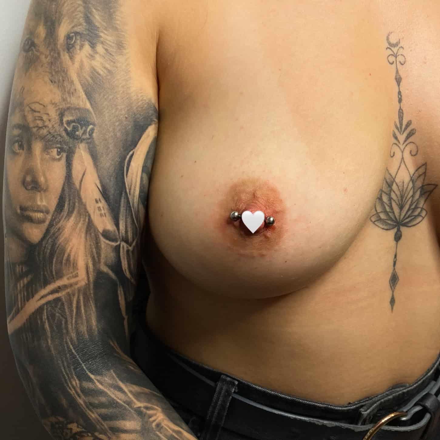 7sinstattoo.pl Osoba z wytatuowanymi rękawami na ramionach, prawdopodobnie ze studia tatuaży Poznań, stoi topless w czarnych spodniach. Ma kolczyk w nosie i piercing w lewym sutku, który jest ozdobiony nakryciem w kształcie serca. Tatuaże mają skomplikowane wzory, w tym portret i obrazy zwierząt. Ten wizerunek jest zgodny z estetyką produktu AF6F23CD-AAA9-42A2-939C-58B01E200F34. Najlepsze studio tatuażu Seven Sins Tattoo