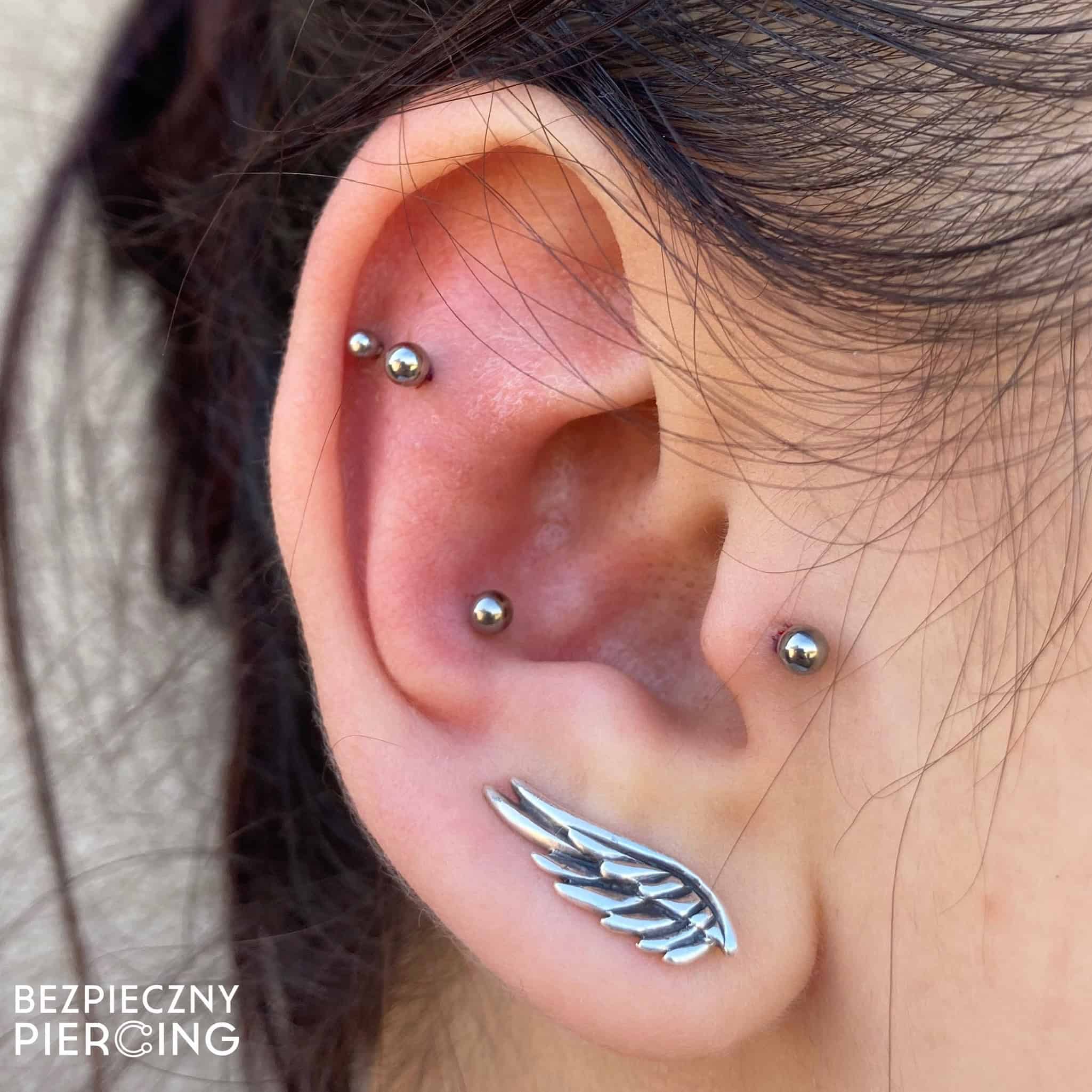 7sinstattoo.pl Zbliżenie ucha zdobionego wieloma piercingami; płatek ucha szczyci się dużym kolczykiem w kształcie skrzydła anioła, a helisa, rook i tragus zdobią małe srebrne kulki. Na zdjęciu znajduje się znak wodny z napisem „Bezpieczny Piercing” pochodzący z renomowanego studia tatuażu Poznań. Najlepsze studio tatuażu Seven Sins Tattoo