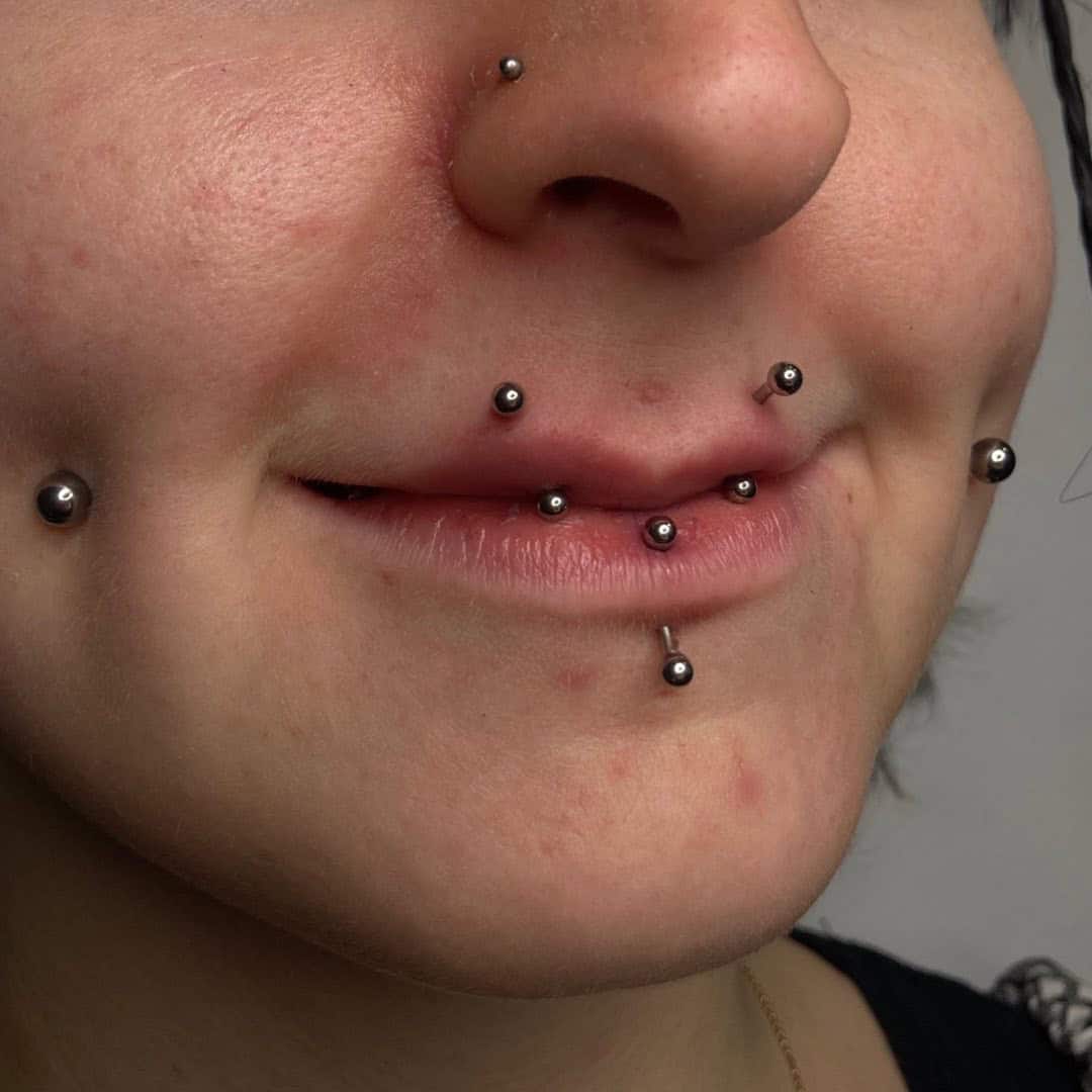 7sinstattoo.pl Zbliżenie dolnej części twarzy osoby pokazujące wiele piercingów twarzy ozdobionych małą metalową biżuterią w kształcie kulek, w tym sześć piercingów na ustach i okolicach, takich jak nos i dołeczki. Osoba ma delikatny uśmiech, pokazując swój unikalny styl z CFEA723B-7BB9-43E0-B620-F338BF248910 z tatuaż Poznań. Najlepsze studio tatuażu Seven Sins Tattoo