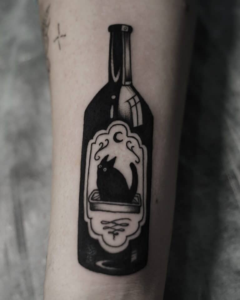 Tatuaż o nazwie DSC00008, pięknie wykonany przez utalentowanych artystów ze studia Seven Sins Tattoo w Poznaniu, przedstawia ciemną etykietę wina z sylwetką wilka wyjącego pod półksiężycem na przedramieniu osoby. Tło jest rozmyte, co wzmacnia elementy dekoracyjne.