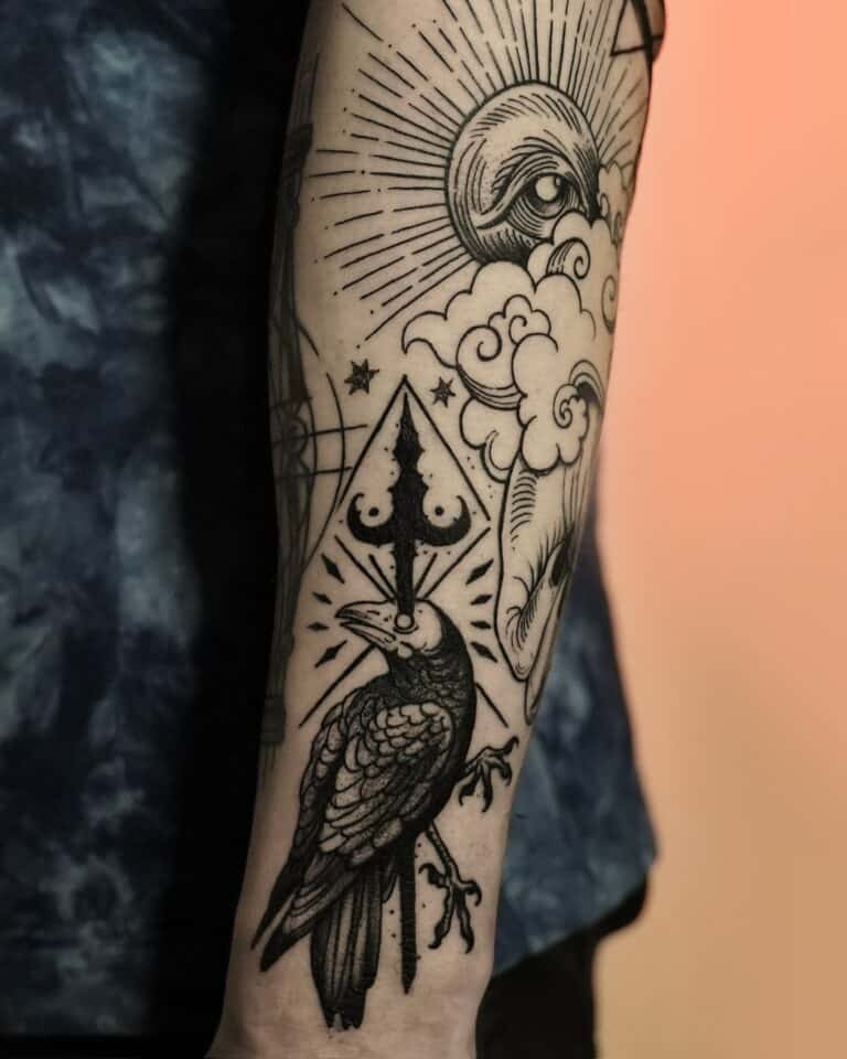 Zbliżenie tatuażu na przedramieniu z kolekcji DSC00032 7sinstattoo.pl, przedstawiającego czarnego kruka siedzącego na gałęzi, z geometrycznym wzorem przypominającym włócznię nad głową. Wokół kruka znajdują się chmury, półksiężyc i skomplikowany wzór linii tworzący wzór oka i rozbłysku słońca. Osoba ma na sobie ciemnoniebieską koszulę.