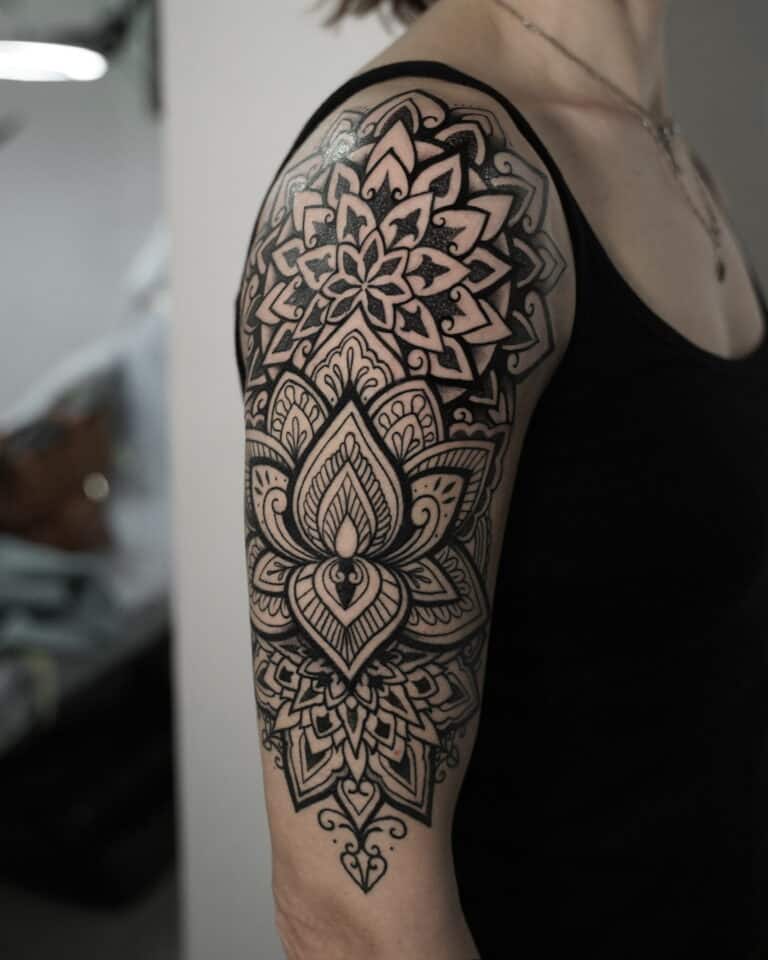 Na zdjęciu widać osobę z krótkimi włosami ubraną w czarny top bez rękawów, z misternym czarnym tatuażem podobnym do tego pokazanego w produkcie DSC00097 z 7sinstattoo.pl. Stworzony w poznańskim studiu Seven Sins Tattoo, ten geometryczny i kwiatowy wzór pokrywa górną część ramienia i bark symetrycznymi wzorami i szczegółowymi motywami mandali, tworząc wizualnie uderzający i wyrafinowany efekt.
