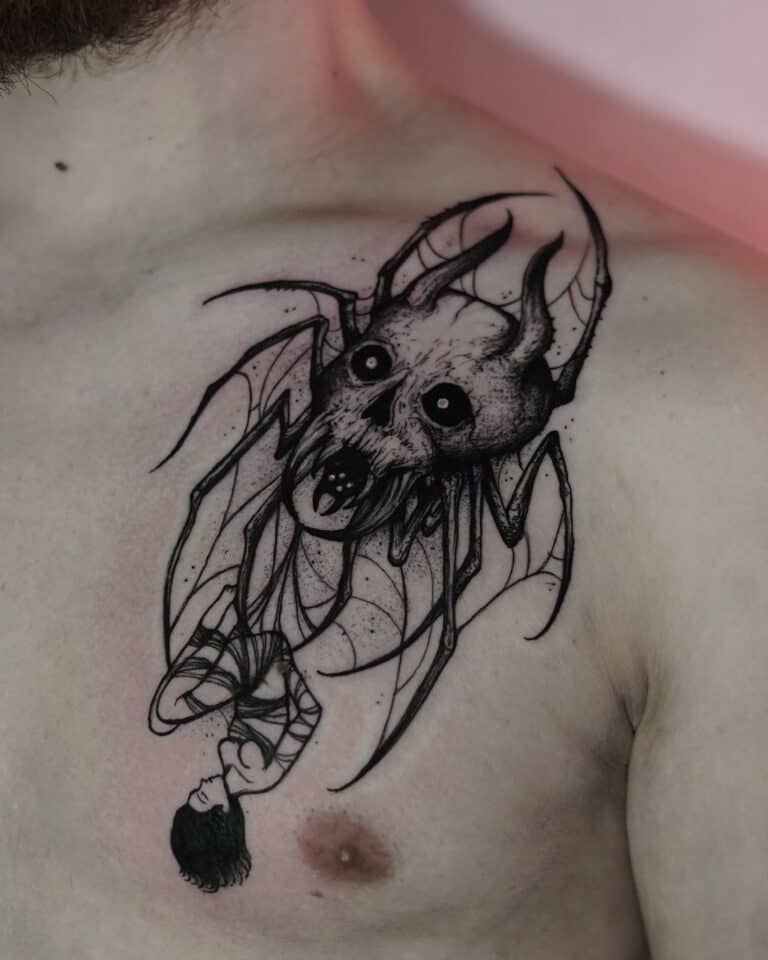 Czarno-biały tatuaż na klatce piersiowej człowieka, misternie wykonany przez renomowane studio 7sinstattoo.pl w Seven Sins Tattoo w Poznaniu, przedstawia groźną czaszkę z nietoperzowymi skrzydłami, kłami i ciemnymi oczami. Wzór obejmuje również wiszącą do góry nogami postać uosabiającą mroczną i gotycką esencję. To arcydzieło uchwycono na zdjęciu DSC00325.