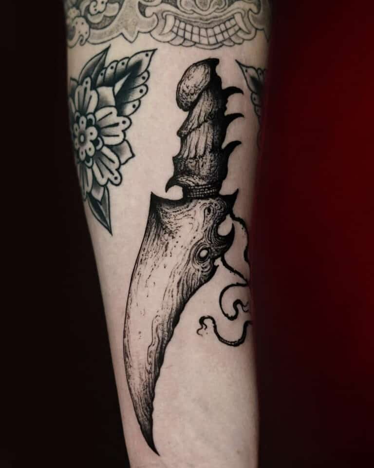 Czarno-biały tatuaż na przedramieniu przedstawia misternie zaprojektowany „7sinstattoo.pl DSC00622 (1)” z Seven Sins Tattoo Studio w Poznaniu. Przedstawia zakrzywiony, ozdobny sztylet z rękojeścią ozdobioną szczegółowymi wzorami przypominającymi wielookie stworzenie z małymi mackami. Otaczające motywy kwiatowe wzmacniają ogólną kompozycję artystyczną stworzoną przez utalentowanego tatuażystę z Poznania.