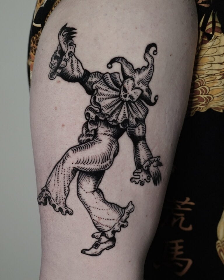 7sinstattoo.pl Zbliżenie na ramię osoby pokazuje szczegółowy czarno-szary tatuaż błazna w klasycznym stroju z falbaniastym kołnierzem, kapeluszem i spiczastymi butami. Błazen jest przedstawiony w animowanej, żywej pozie, emanującej kaprysem. Uchwycony jako DSC04234 w tatuaż Poznań, osoba ma na sobie ciemne, wzorzyste ubranie. Najlepsze studio tatuażu Seven Sins Tattoo