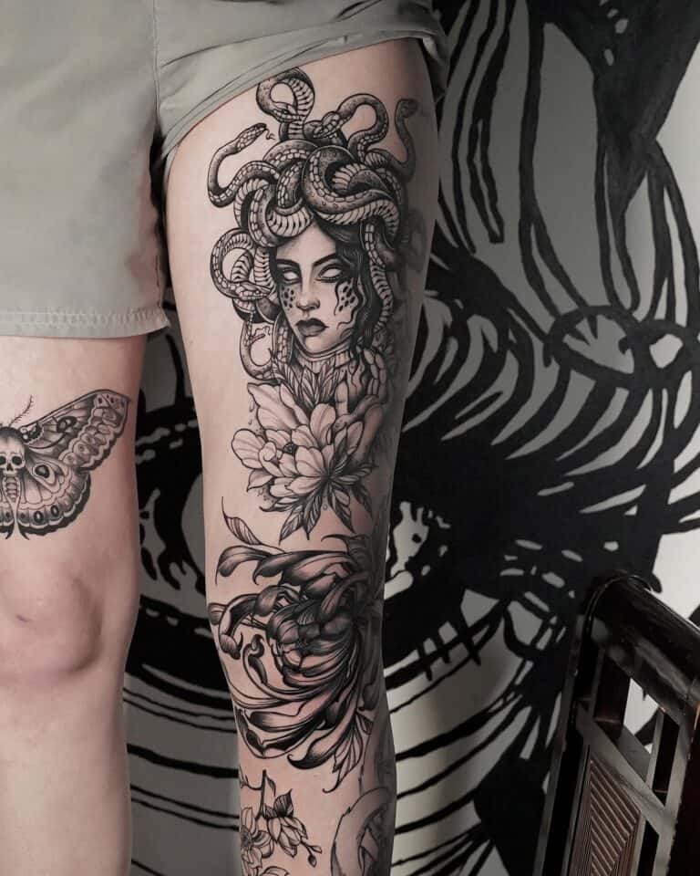 7sinstattoo.pl Wytatuowana noga z dużą głową Meduzy z włosem węża, różą i innymi skomplikowanymi wzorami, takimi jak ćma i krab, rozciąga się od połowy uda do kolana. Szczegółowe monochromatyczne dzieło sztuki wyróżnia się na tle czarnego muralu, wszystko fachowo wykonane w studiu DSC04304-восстановлено w Poznaniu. Najlepsze studio tatuażu Seven Sins Tattoo