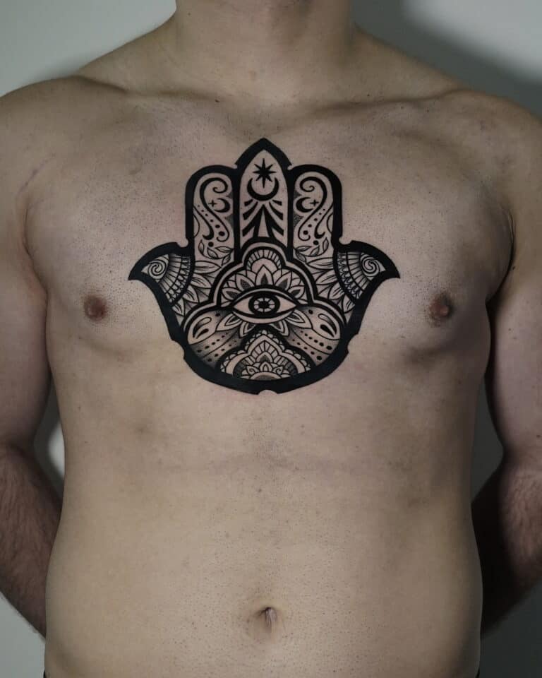 7sinstattoo.pl Osoba z dużym, misternym czarnym tatuażem symbolu Hamsa na piersi stoi na prostym tle. Wzór charakteryzuje się ozdobnymi wzorami i obejmuje oko w środku dłoni, rozciągające się na górną część klatki piersiowej. Ta oszałamiająca praca została uchwycona w produkcie DSC04471 w studio tatuażu Poznań. Najlepsze studio tatuażu Seven Sins Tattoo