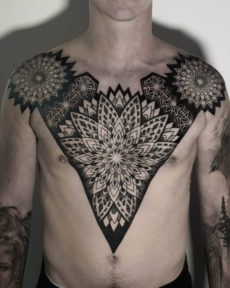 7sinstattoo.pl Osoba z misternymi czarnymi geometrycznymi tatuażami pokrywającymi klatkę piersiową i ramiona, które zostały wykonane w najlepszym studio tatuażu w Poznaniu. Centralny wzór przedstawia duży, symetryczny wzór kwiatowy, podczas gdy mniejsze sąsiadujące wzory uzupełniają go na ramionach. Ta szczegółowa i symetryczna sztuka ciała została uchwycona w produkcie DSC04820. Najlepsze studio tatuażu Seven Sins Tattoo