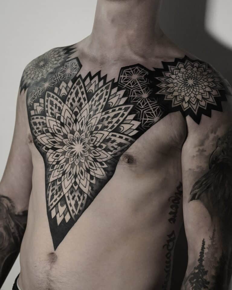 7sinstattoo.pl Osoba prezentuje misterny i rozległy czarno-szary tatuaż mandali, który rozciąga się na klatce piersiowej i ramionach, z dodatkowymi mniejszymi tatuażami zdobiącymi jej ramię. Oświetlenie podkreśla szczegółowy projekt i cieniowanie widocznego tatuażu na klatce piersiowej, umiejętnie wykonanego w tatuaż Poznań, jak uchwycono w produkcie DSC04850. Najlepsze studio tatuażu Seven Sins Tattoo