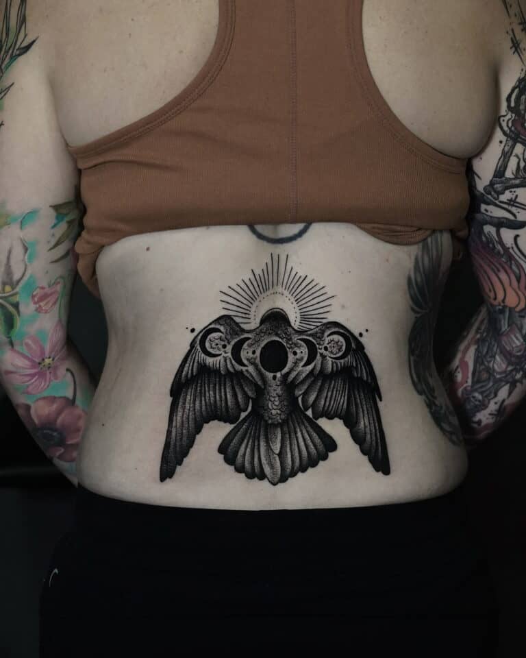 7sinstattoo.pl Osoba oznaczona jako DSC05694 ma tatuaż na plecach przedstawiający szczegółowego, ciemno cieniowanego ptaka z rozpostartymi skrzydłami, okrągłym wzorem przypominającym słońce nad głową i misterną kropką. Ma na sobie brązowy krótki top i ciemne spodnie, odsłaniając kolorowe tatuaże na ramionach wykonane w najlepszym studiu tatuażu Poznań. Najlepsze studio tatuażu Seven Sins Tattoo