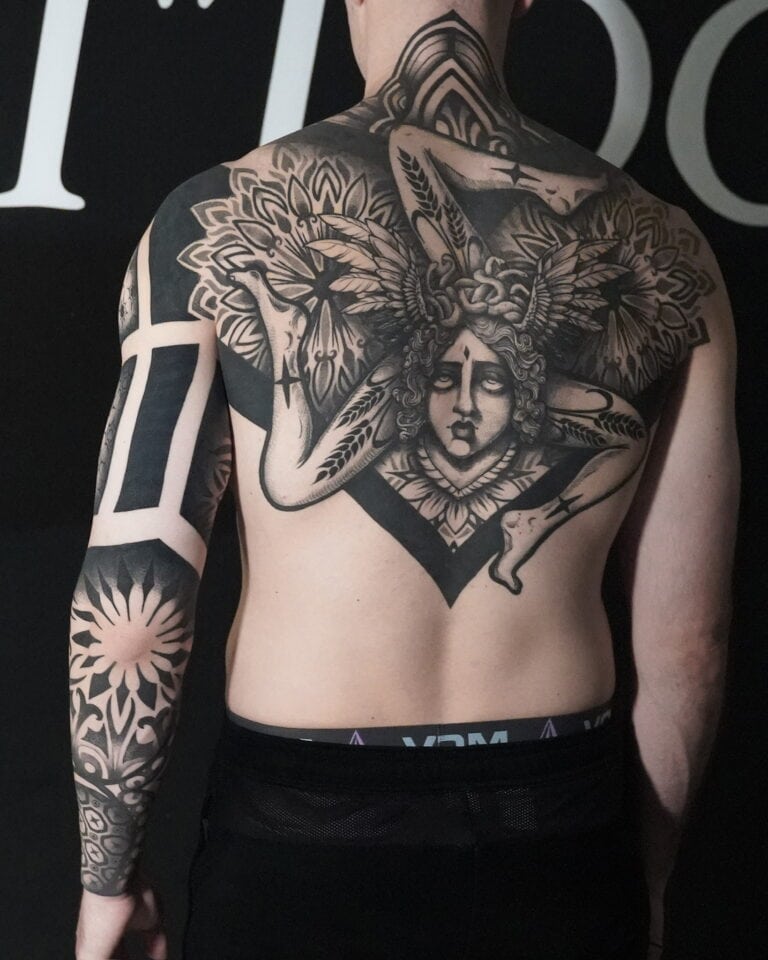 7sinstattoo.pl Na plecach osoby znajduje się szczegółowy czarno-szary tatuaż z geometrycznym wzorem i postacią z misternymi skrzydłami oraz ozdobnym nakryciem głowy, stworzony w najlepszym studio tatuażu w Poznaniu. Osoba ta ma również abstrakcyjne i kwiatowe tatuaże na prawym ramieniu. Więcej szczegółów można znaleźć w nazwie produktu: DSC05897. Najlepsze studio tatuażu Seven Sins Tattoo