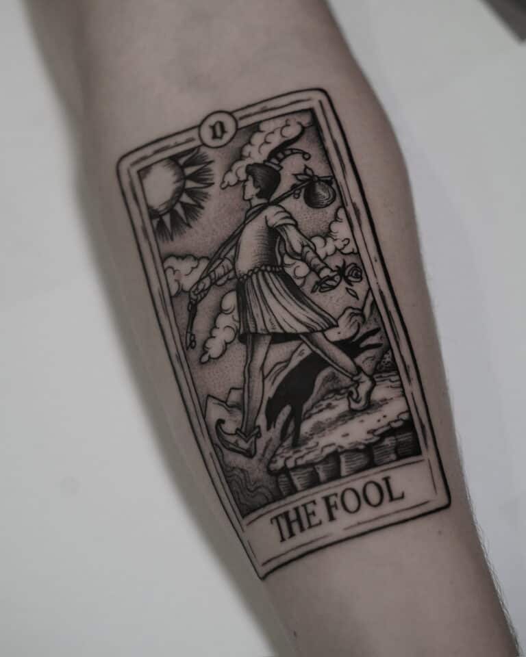 7sinstattoo.pl Szczegółowy czarno-biały tatuaż karty Tarota „Głupiec” na przedramieniu, uchwycony na zdjęciu produktu DSC09590 z najlepszego studia tatuażu Poznań. Wzór przedstawia osobę w stroju błazna schodzącą z klifu, trzymającą mały woreczek na kiju na ramieniu, ze słońcem i chmurami w tle. Najlepsze studio tatuażu Seven Sins Tattoo
