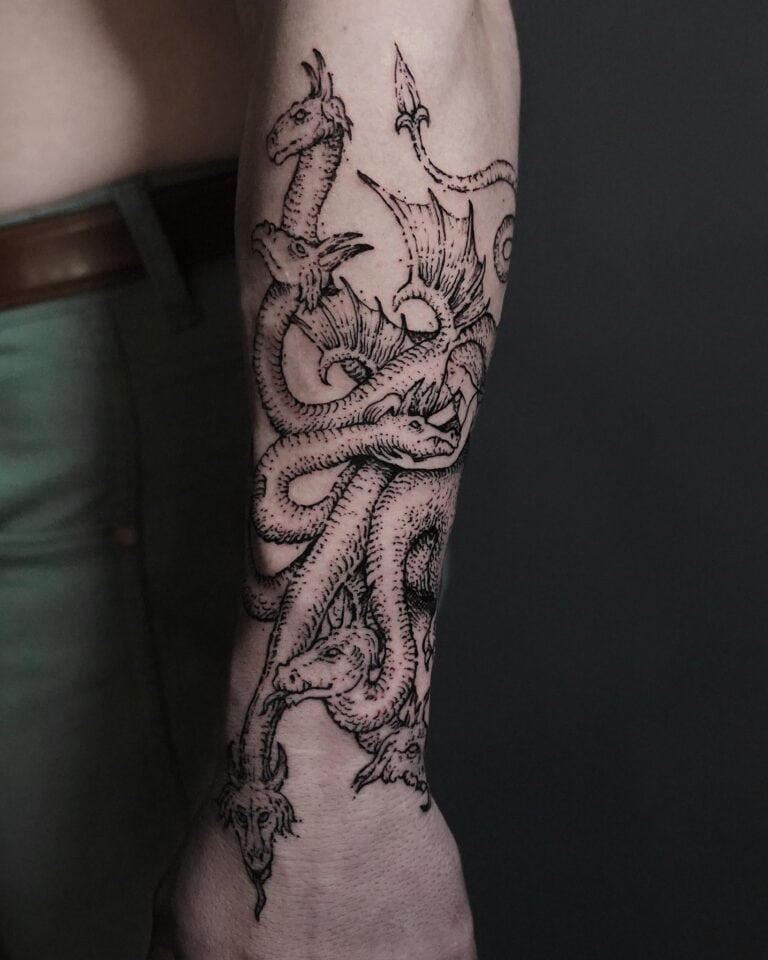 7sinstattoo.pl Zbliżenie na przedramię osoby, na którym widoczny jest skomplikowany tatuaż wielogłowego smoka, wykonany przez utalentowanego artystę z najlepszego studia tatuażu w Poznaniu. Szczegółowa praca liniowa ukazuje głowy smoków warczące i splatające się wzdłuż przedramienia. Osoba, zidentyfikowana jako DSC09812, ma na sobie zielone spodnie. Najlepsze studio tatuażu Seven Sins Tattoo