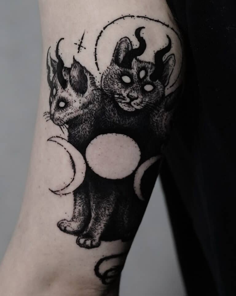 7sinstattoo.pl Oszałamiający tatuaż zatytułowany „DSC09934”, stworzony w najlepszym studio tatuażu Poznań, przedstawia dwugłowego kota z trzema oczami i rogami. Obie głowy mistycznego stworzenia mają świecące oczy i są otoczone elementami niebieskimi, w tym fazami księżyca. Ta czarno-biała grafika zdobi ramię osoby na ciemnym tle. Najlepsze studio tatuażu Seven Sins Tattoo