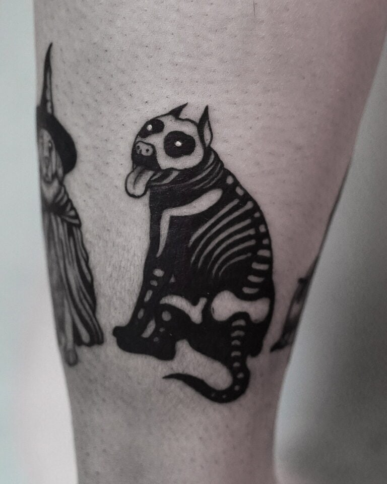 7sinstattoo.pl Tatuaż na nodze osoby przedstawiający czarno-białego stylizowanego psa o szkieletowych rysach i wystającym języku, obok częściowego obrazu postaci w szacie i kapeluszu. Projekt utrzymuje mroczny, kapryśny motyw i został wykonany przez artystę z najlepszego studia tatuażu Poznań. Ta praca jest identyfikowana jako DSC09947. Najlepsze studio tatuażu Seven Sins Tattoo