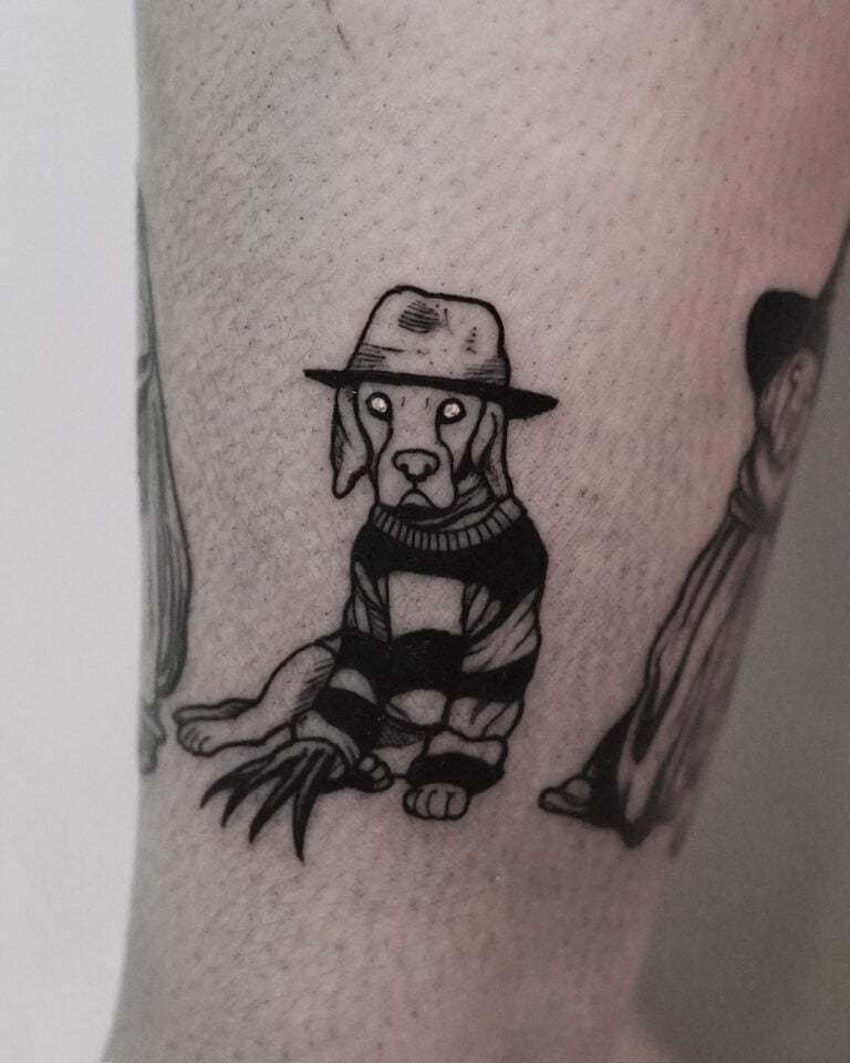 7sinstattoo.pl Czarno-biały tatuaż, wykonany w studio tatuaży Poznań, przedstawia psa w szerokim kapeluszu i pasiastym swetrze, przypominającym postać z horroru. Pies ma pazury w rękawiczkach na jednej łapie, co dodaje niepokojącego motywu, i siedzi na jasnym ramieniu osoby. Tatuaż jest skatalogowany jako produkt DSC09972. Najlepsze studio tatuażu Seven Sins Tattoo